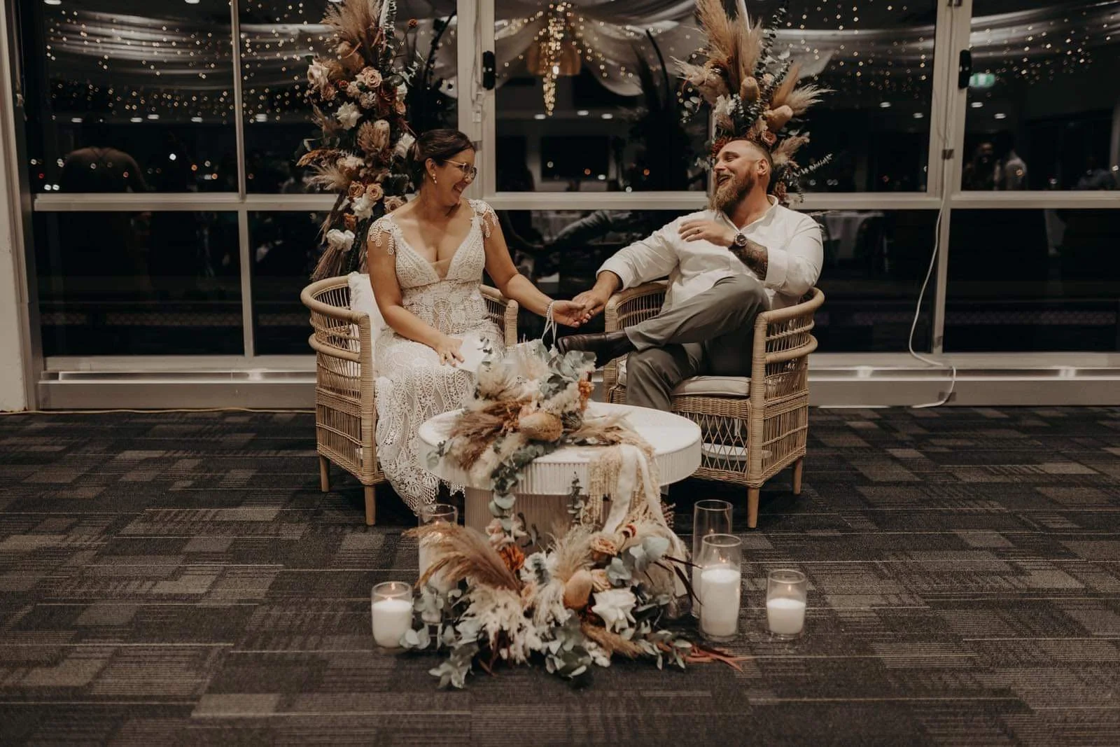 A bride and groom sitting on wicker chairs, holding hands and smiling at each other during their wedding reception at night, with floral arrangements and candles on the floor