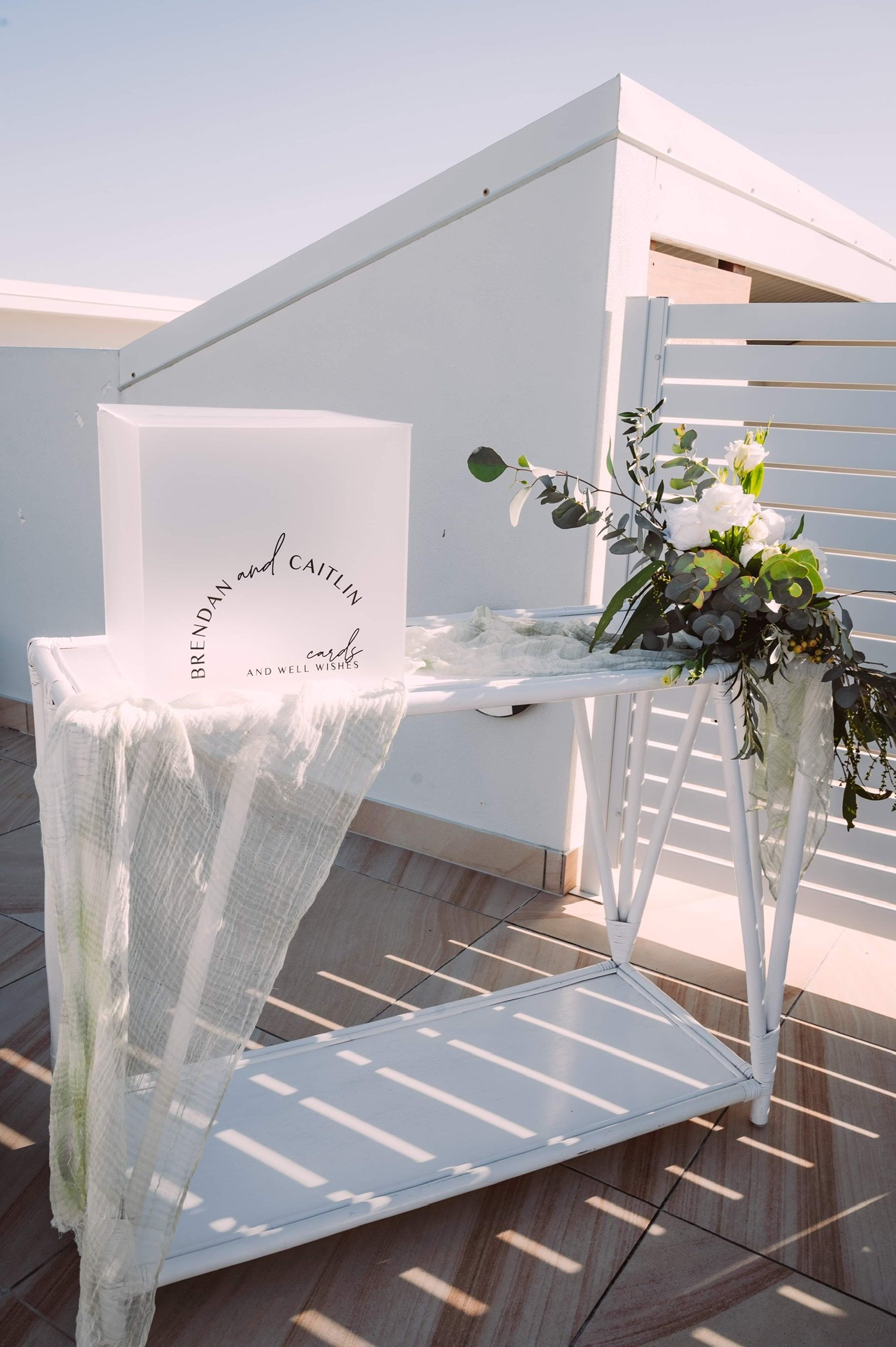 Wedding card display with a white wedding card and a floral arrangement on a white table, outdoors with sunny weather.