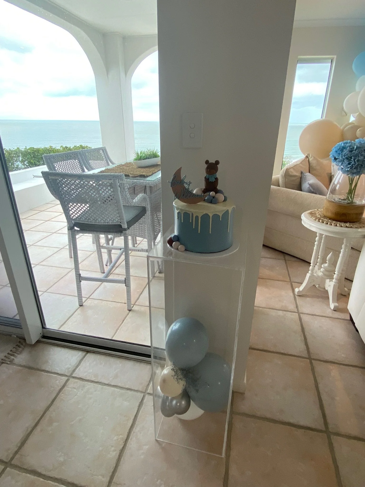 Decorative cake with teddy bear topper and blue, white, and red balls inside a clear acrylic container, with a view of an ocean balcony in the background.