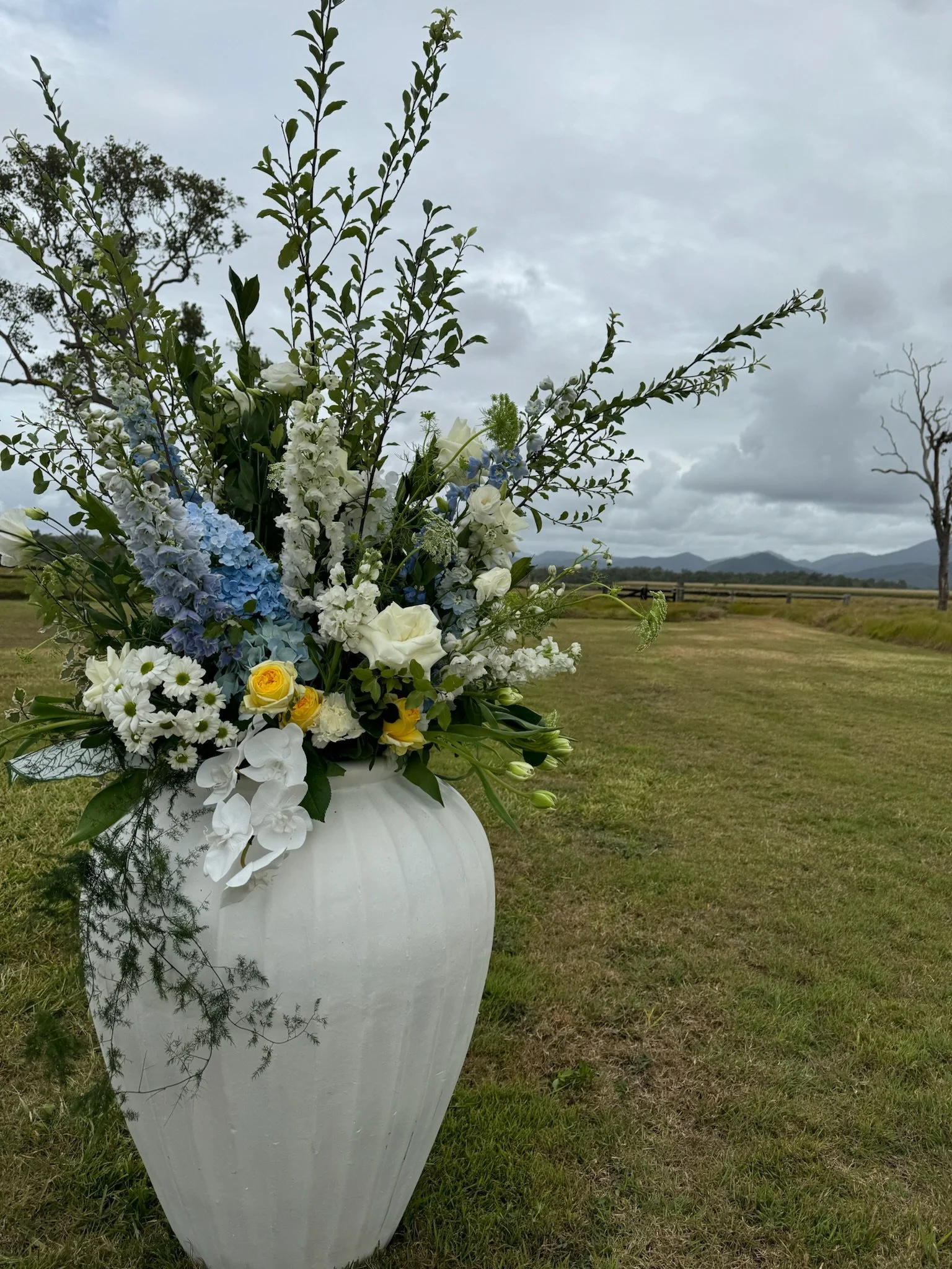 A large white vase filled with a variety of white, yellow, and blue flowers, positioned outdoors on a grassy field with cloudy sky and distant mountains in the background.