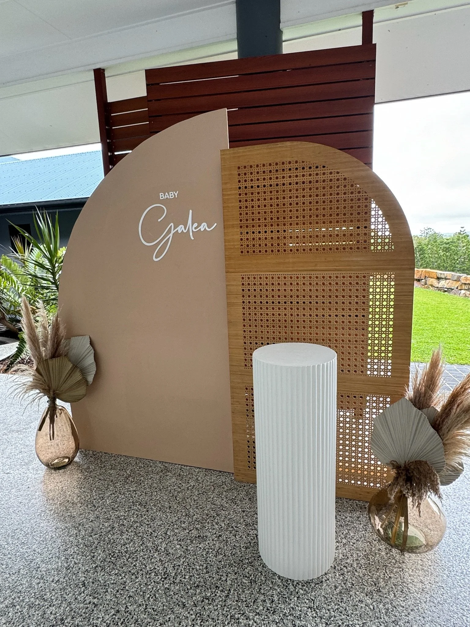 Decorative baby shower display with beige and wooden panels, vases with dried pampas grass, and a white cylindrical stand, outdoors with green grass and cloudy sky in the background.