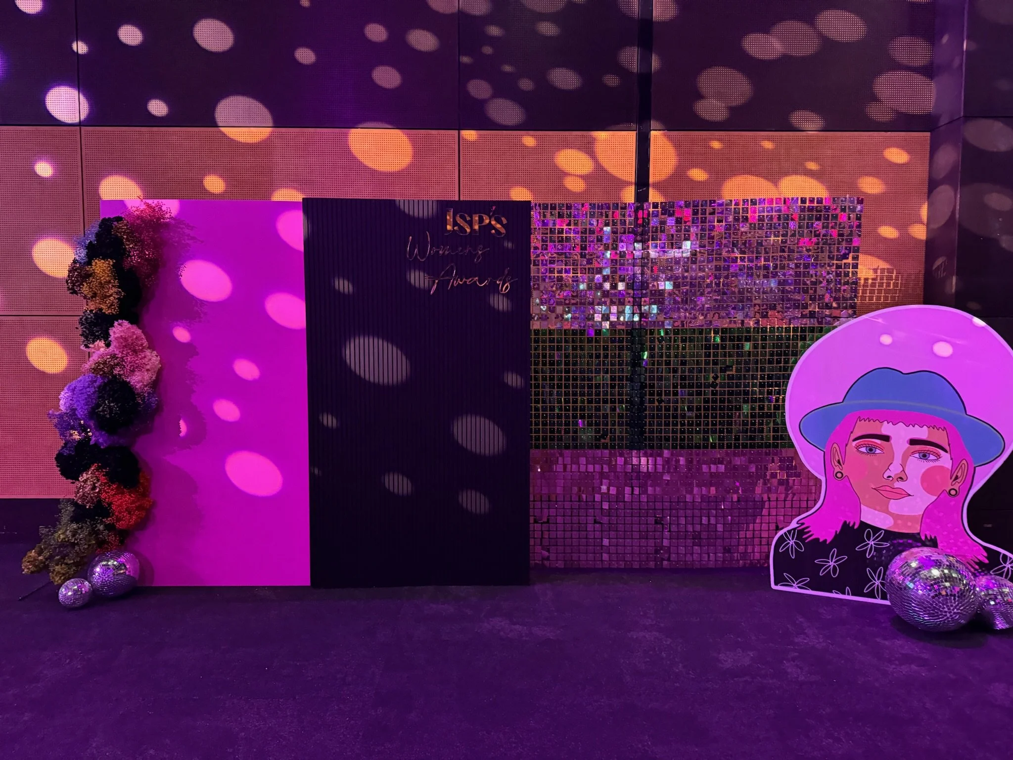 Decorated backdrop for the ISPS Women's Awards event with colorful flowers, reflective disco balls, and a large illustrated portrait of a woman wearing a hat, all illuminated by purple lighting.
