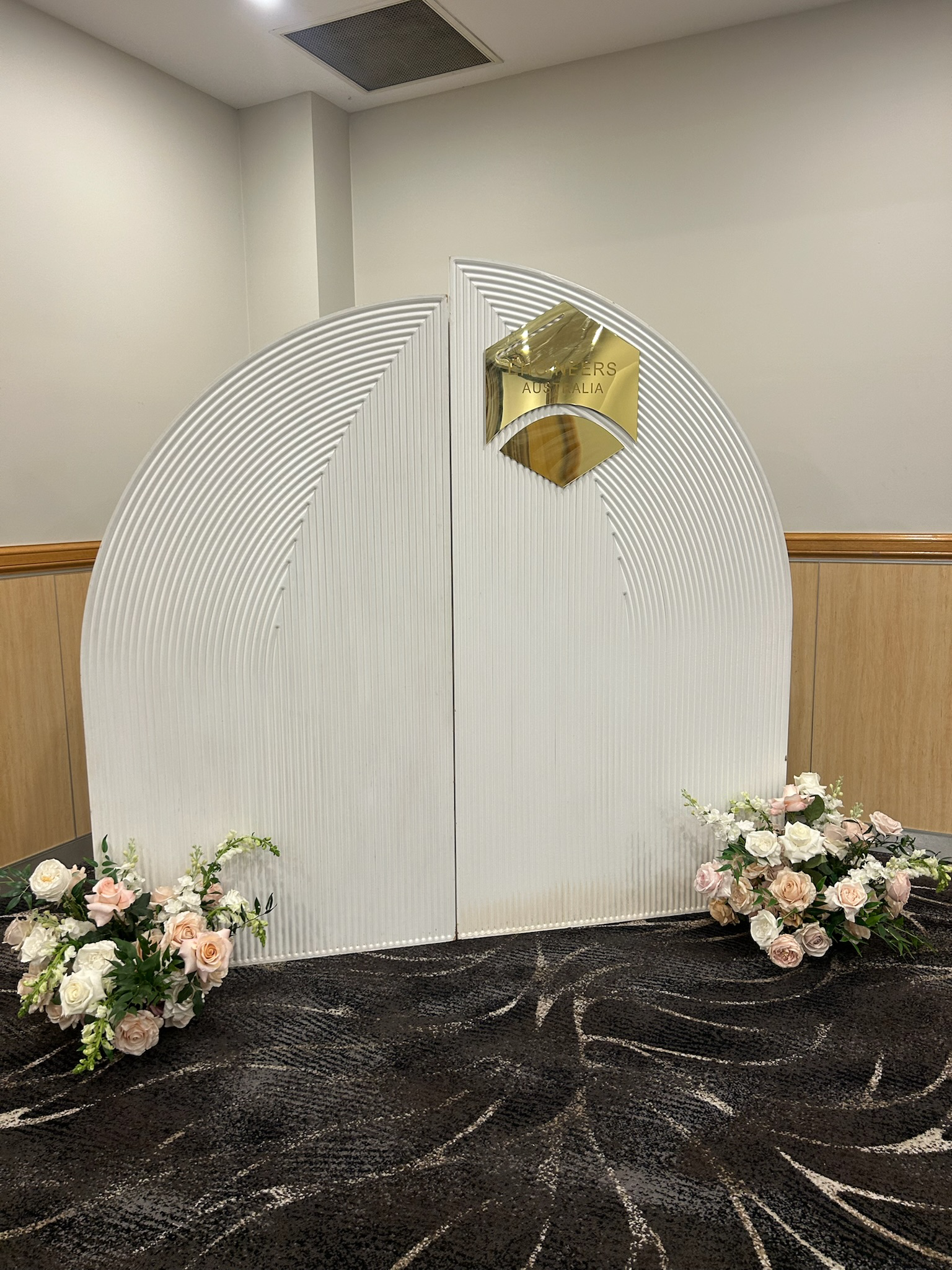 A decorative backdrop with white curved panels, adorned with pink and white floral arrangements on a dark patterned carpet.