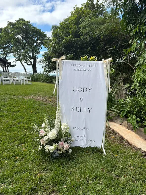 White wedding sign with welcome message for Cody and Kelly, dated May 17, 2023, located outdoors on green grass with trees and chairs in the background, decorated with white flowers and ribbons.