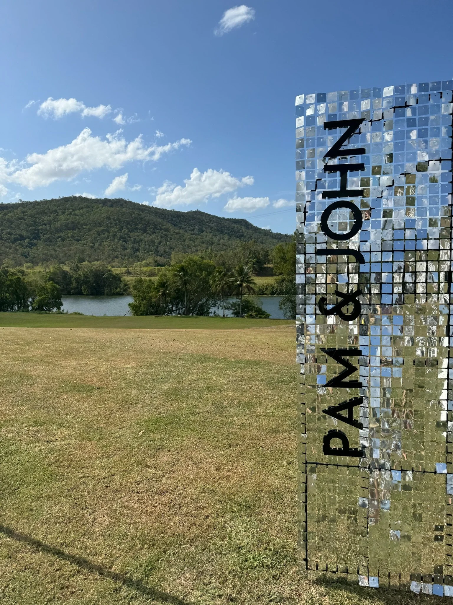 Reflection of trees and sky on a large, mirrored sign that reads 'PAWAN & JOHN' in black letters, on a grassy field with a lake, palm trees, and a mountain in the background under a partly cloudy sky.
