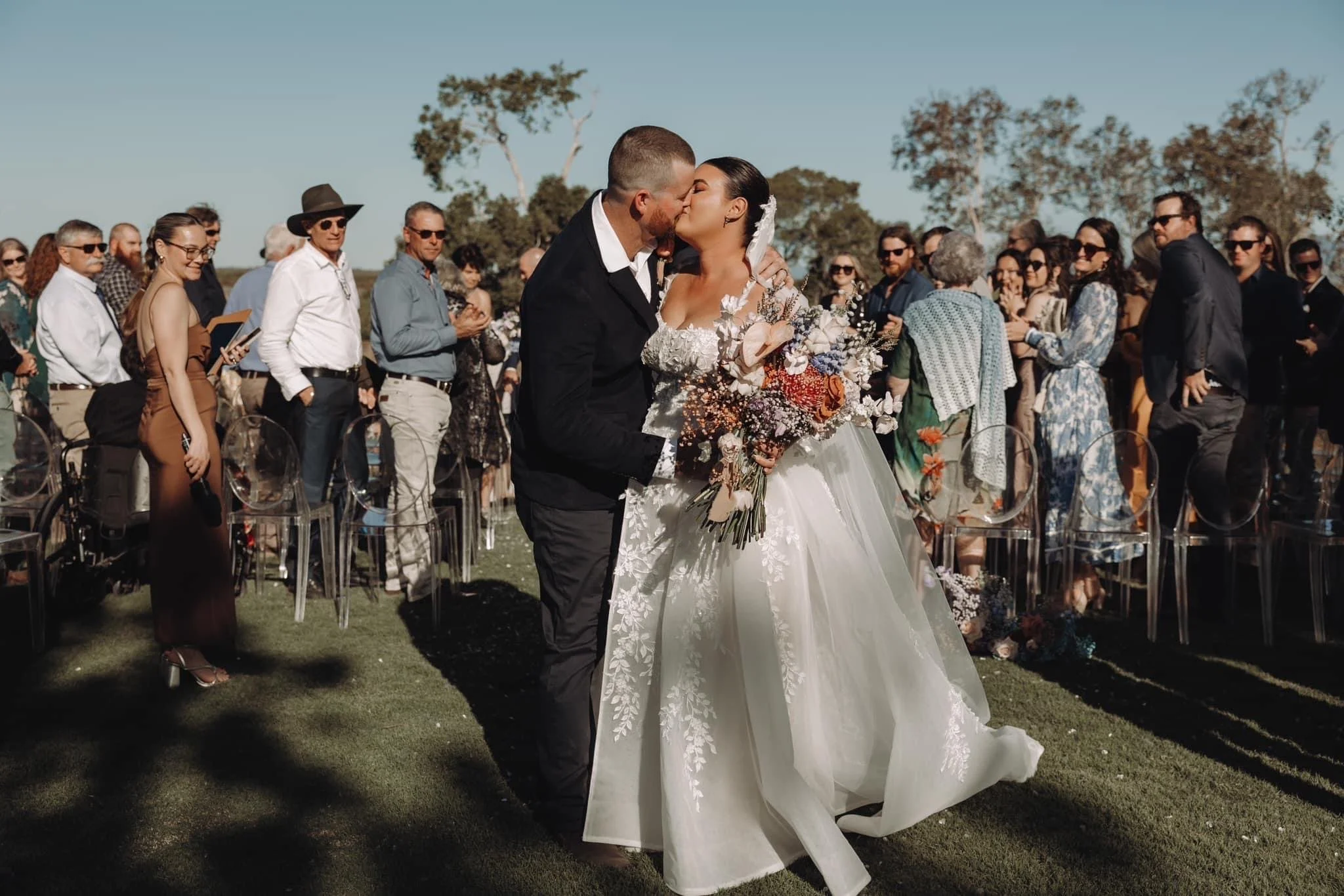 A bride and groom kiss during their outdoor wedding ceremony, surrounded by seated guests and tall trees under a clear sky.