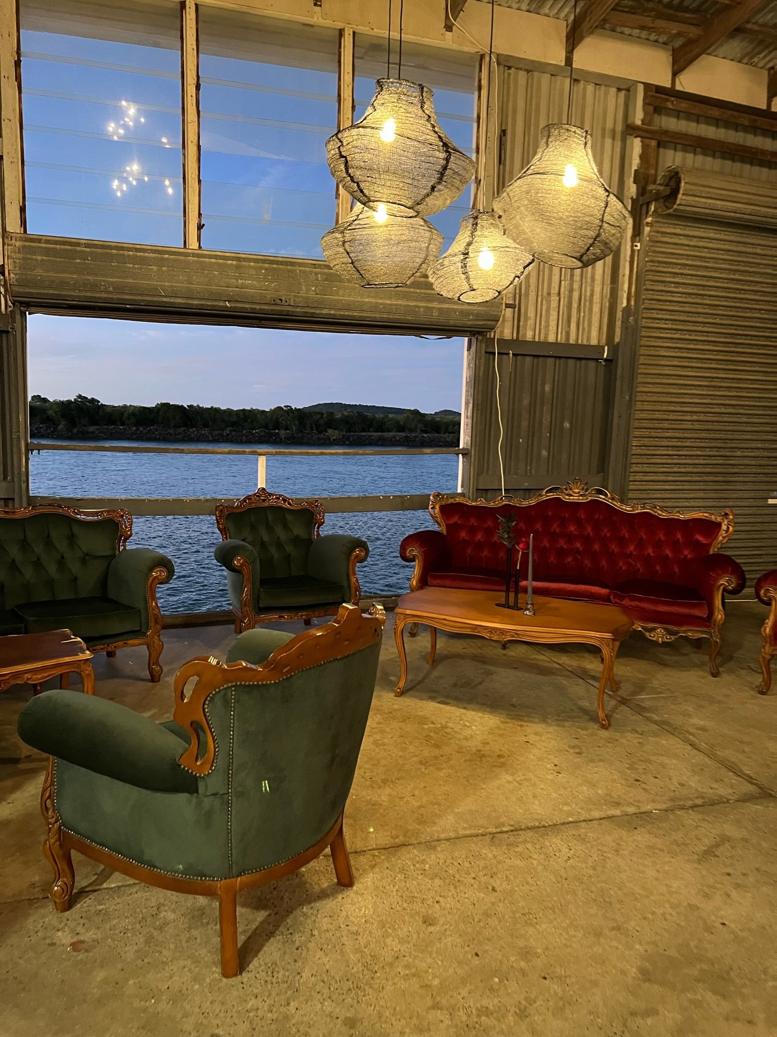 Indoor seating area with vintage-style green and red velvet couches and armchairs, wooden coffee table, and unique hanging light fixtures. Large window shows a view of a body of water and distant land at dusk.