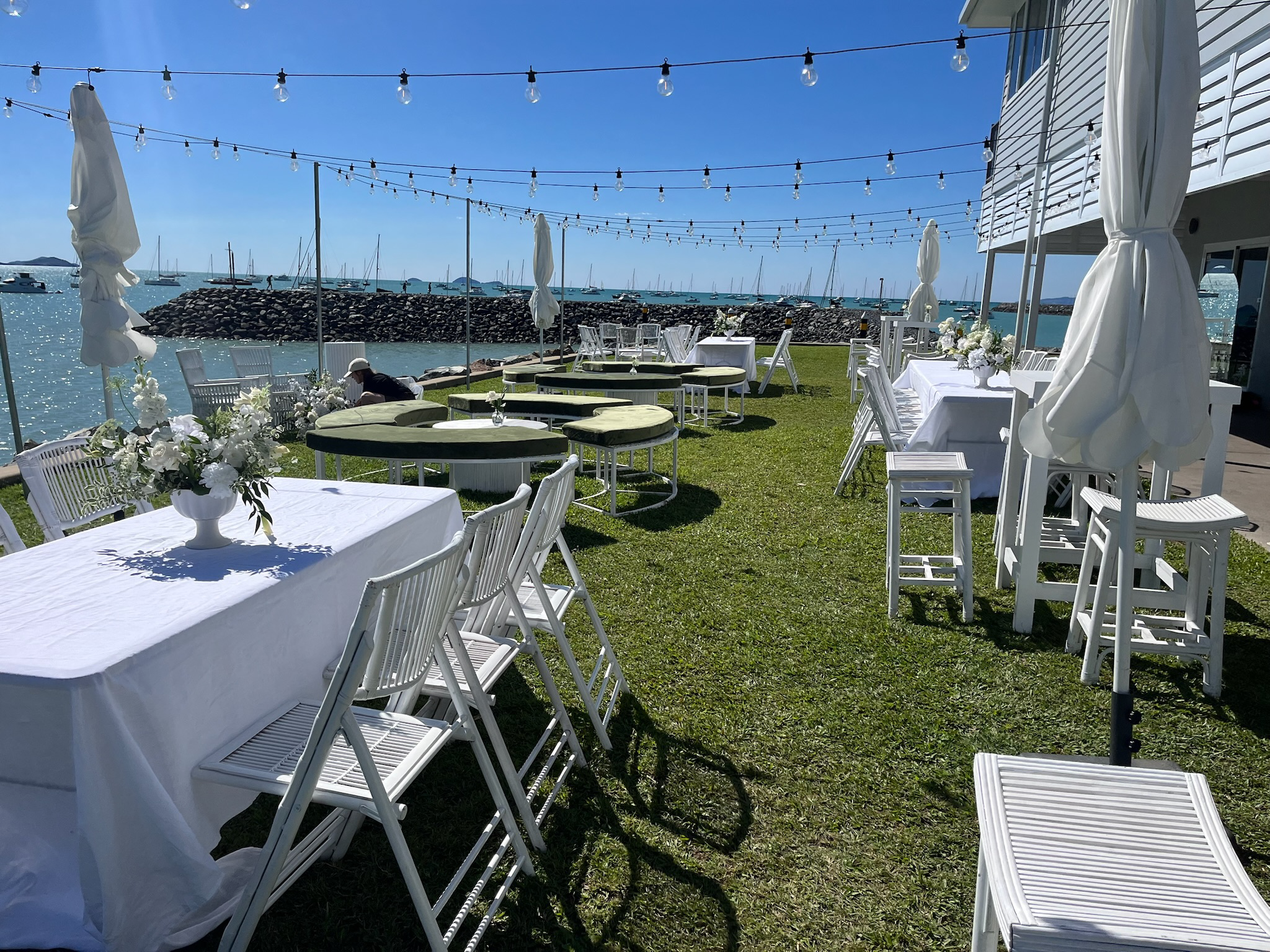 Outdoor dining area with white tables and chairs, decorated with white flowers, near the water with sailboats in the distance, string lights overhead, and umbrellas, on a sunny day.