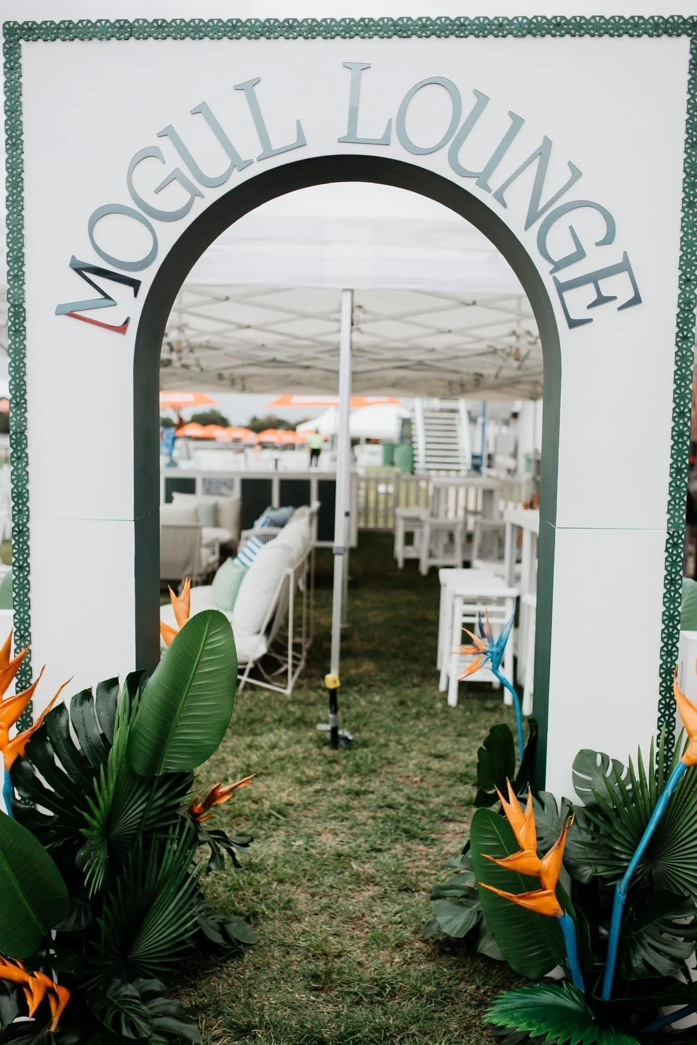 Arched entrance with 'MOUCHALL LOUNGE' sign, tropical plants and flowers framing the entrance, outdoor seating area with white furniture and umbrellas in background.