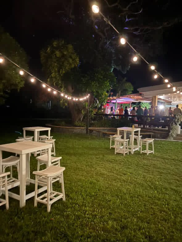 Outdoor event space at night with string lights, green grass, white tables and stools, and a building with people inside.