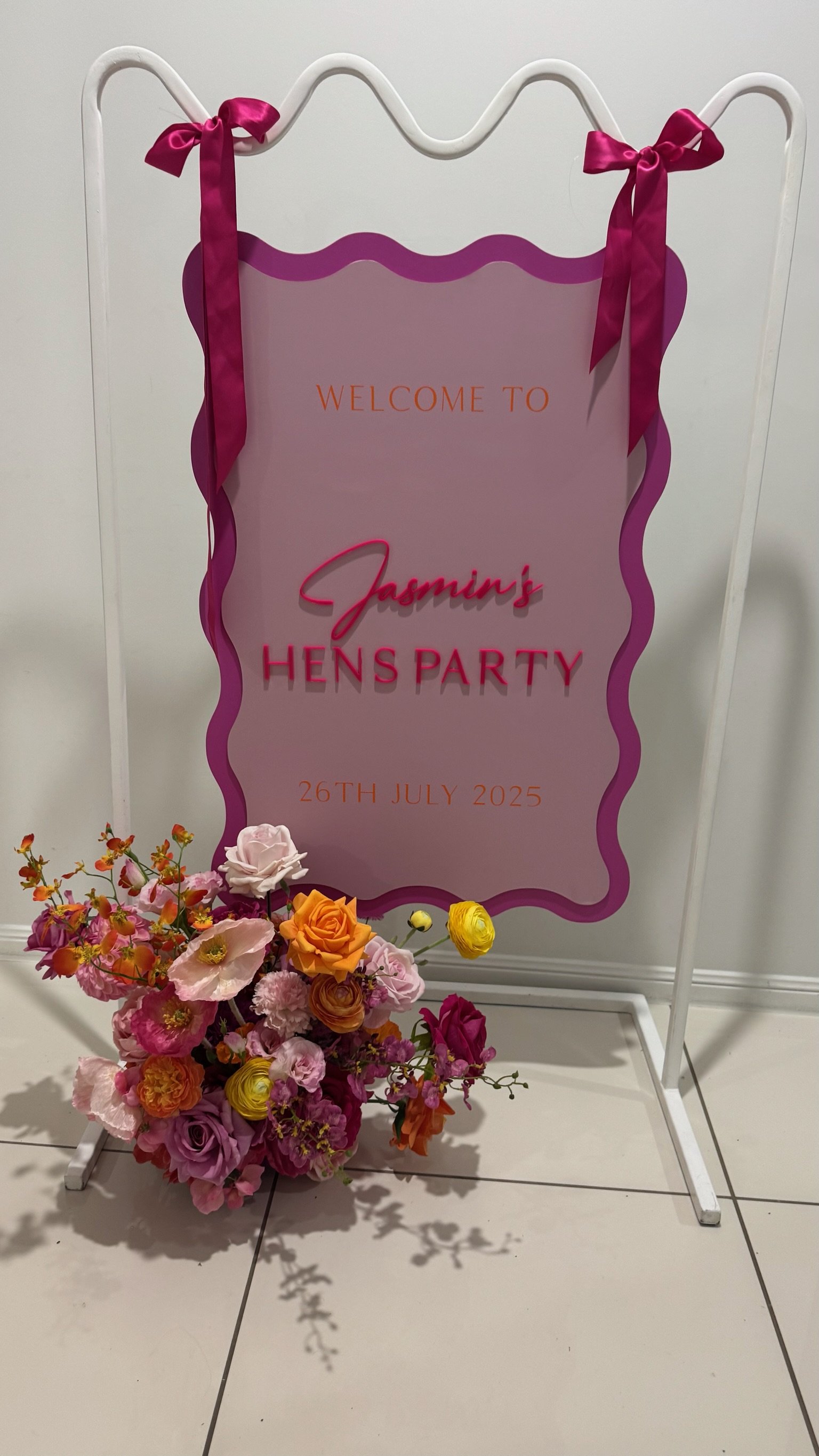 A welcome sign for Jasmine's Hens Party, dated July 26, 2025, with a pink and purple design, decorated with pink ribbons and a bouquet of pink, orange, purple, and yellow flowers in front.