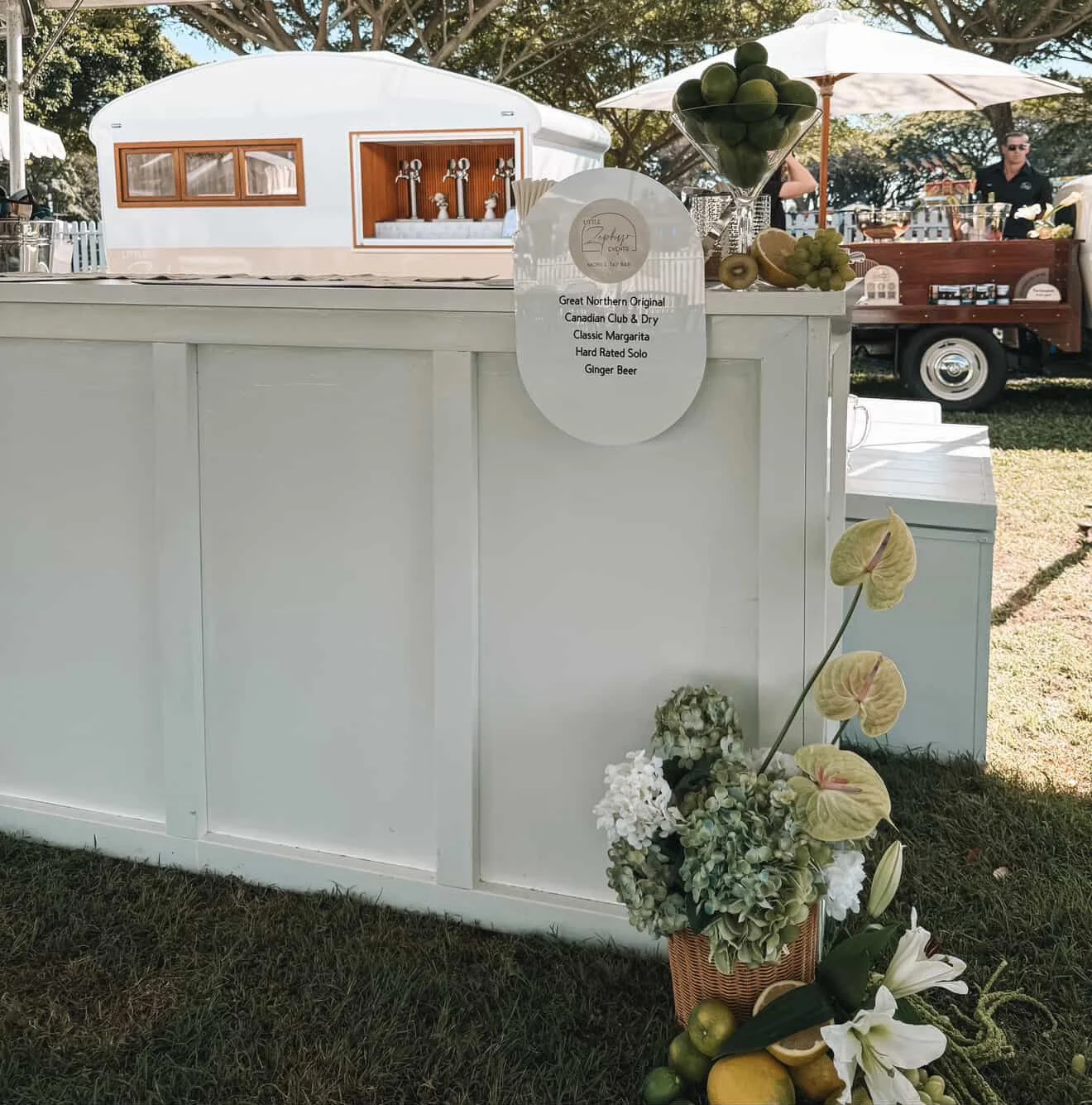 A white outdoor bar with a sign listing drink options, decorated with flowers and fruits, with a small trailer and people in the background.