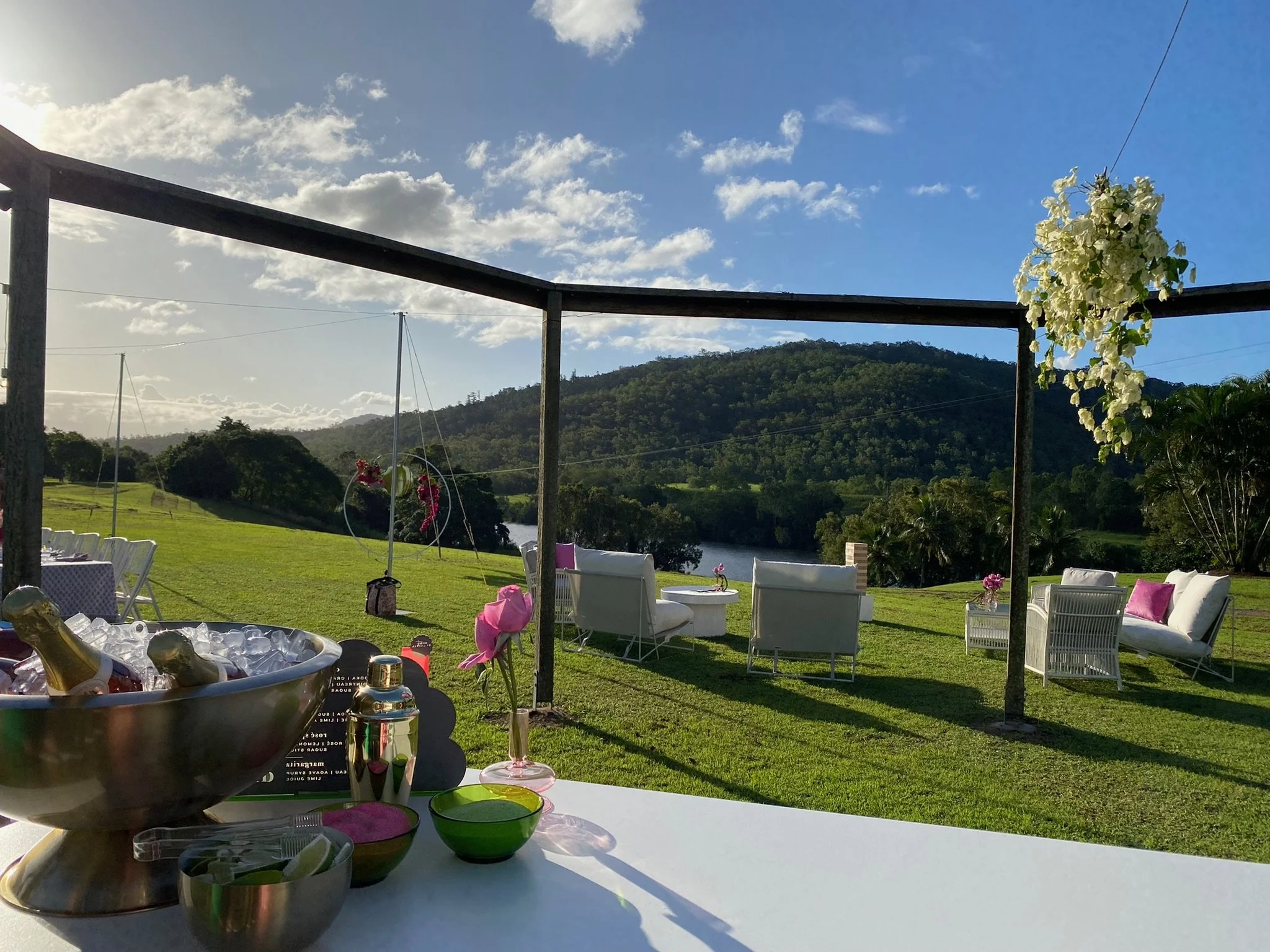 Outdoor setting with white chairs and sofas, pink cushions, decorated flowers, and a scenic view of green hills, trees, a river, with a partly cloudy sky.