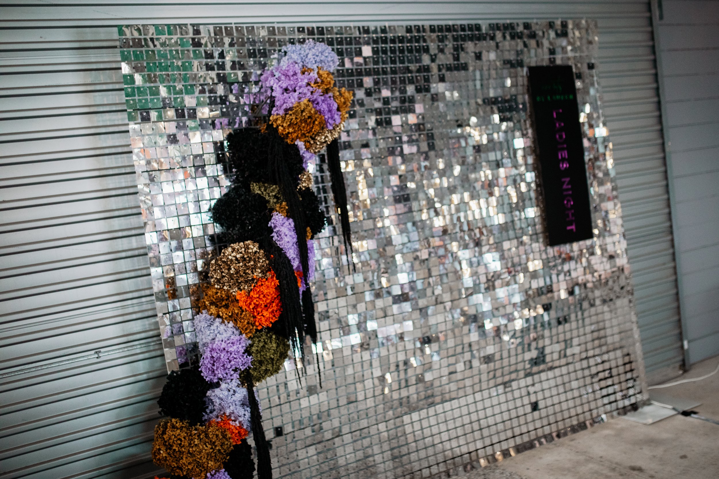 A decorative display with a mosaic tile backdrop and a floral arrangement with purple, orange, and black flowers, along with a sign that reads 'LADIES NIGHT' in purple and pink letters.