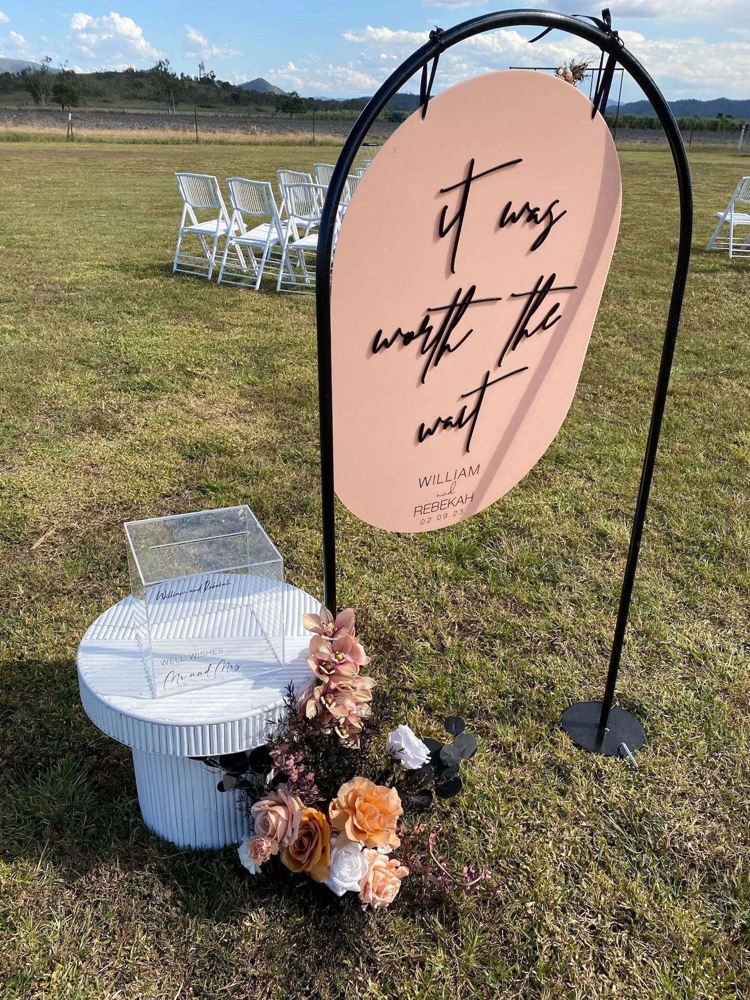 Outdoor wedding decoration with a large pink sign that says 'it was worth the wait' and includes names William and Rebekah with the date 02.09.23. A small table with wedding wishes and a floral arrangement is nearby. White chairs are arranged in the 