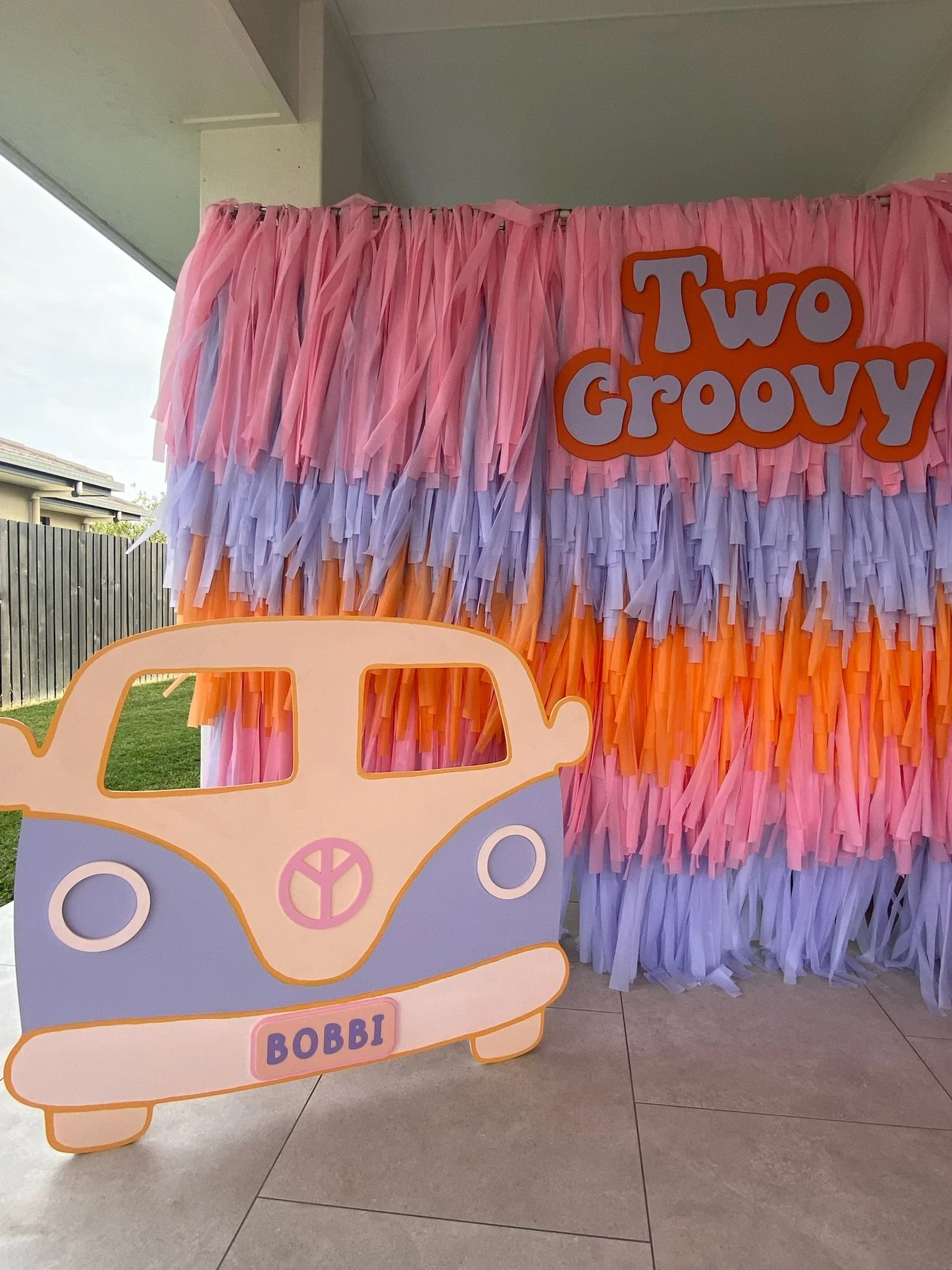 Decorative background with pink, purple, and orange tissue paper tassels, red and white 'Two Groovy' sign, and a cutout of a vintage Volkswagen van with a peace sign on the front and a custom license plate that says 'BOBBI'.