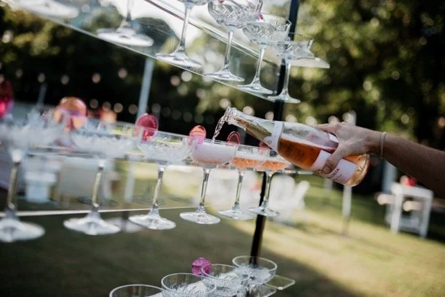 Person pouring a bottle of beer into a champagne glass structure made of multiple tiers of glasses at an outdoor event.