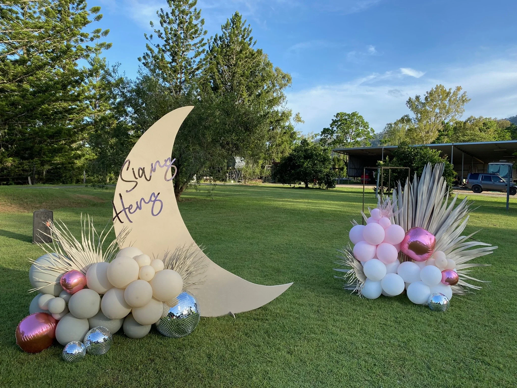 A decorative setup with a large cream-colored crescent moon cutout with the name 'Siary Henz' written on it, surrounded by clusters of white, pink, and metallic balloons, and dried plant arrangements, outdoors on a grassy field with trees and a cover