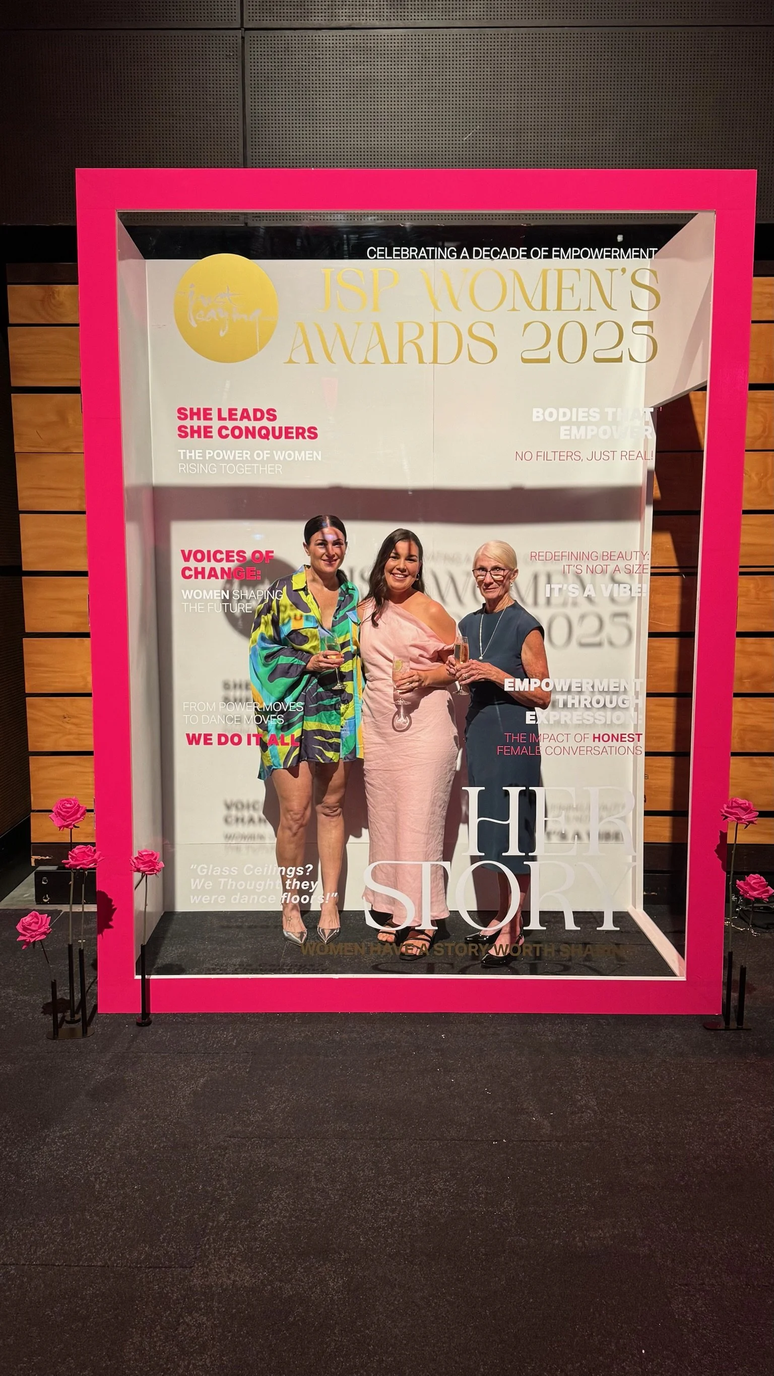 Three women standing together inside a decorative photo frame at the JSP Women's Awards 2025 event, smiling and holding champagne glasses, with event posters and pink roses around them.