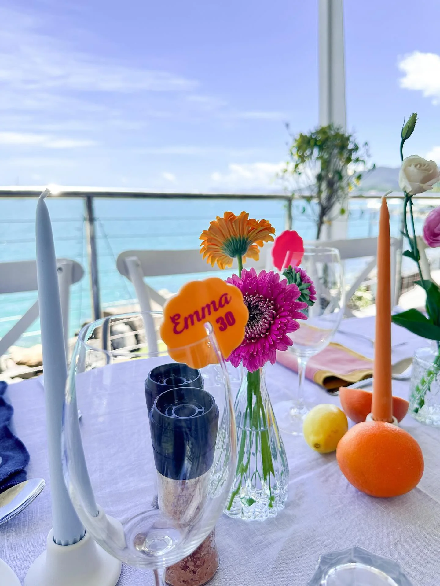 A decorated table with colorful flowers in a glass vase, fruit, candles, and place settings, overlooking a body of water and a blue sky.