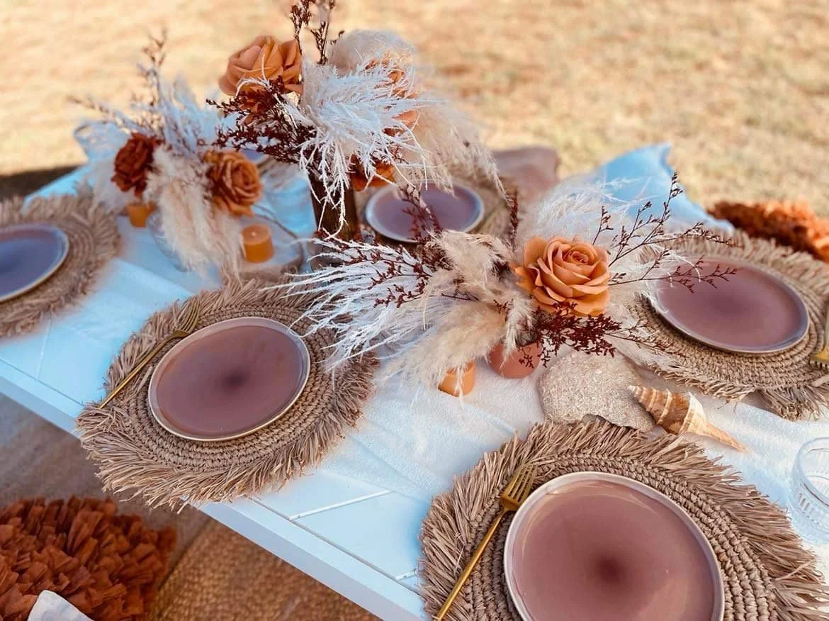 Table setting with brown woven placemats, pink plates, gold flatware, and a centerpiece with orange roses, dried white and brown foliage, and seashells on a white tablecloth outdoors.