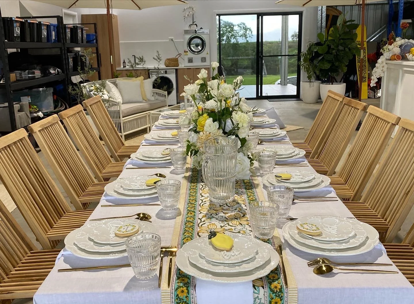 A long dining table set for a meal with white plates, gold utensils, and glassware, decorated with a floral table runner and a large bouquet of white and yellow flowers in the center, in a bright room with a sliding glass door and outdoor view.