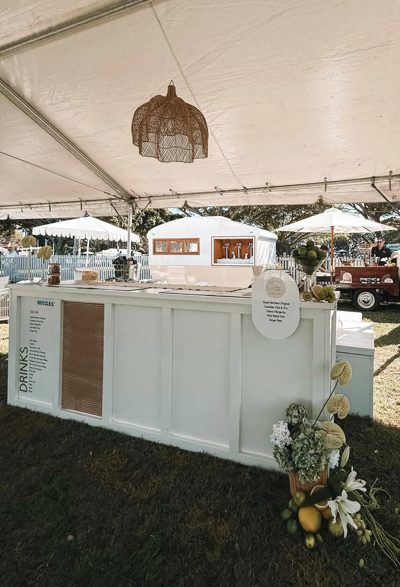 White outdoor refreshment stand with a sign listing drinks, decorated with flowers and fruit, located under a beige tent at an outdoor event.