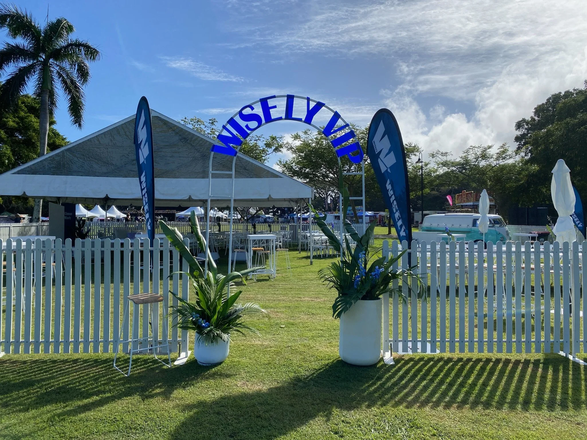 Outdoor event setup with a white picket fence, a large white tent, blue inflatable flags, and a sign that reads 'Misely VIP' in blue, with green plants in white pots, on a grassy area under a partly cloudy sky with palm trees in the background.