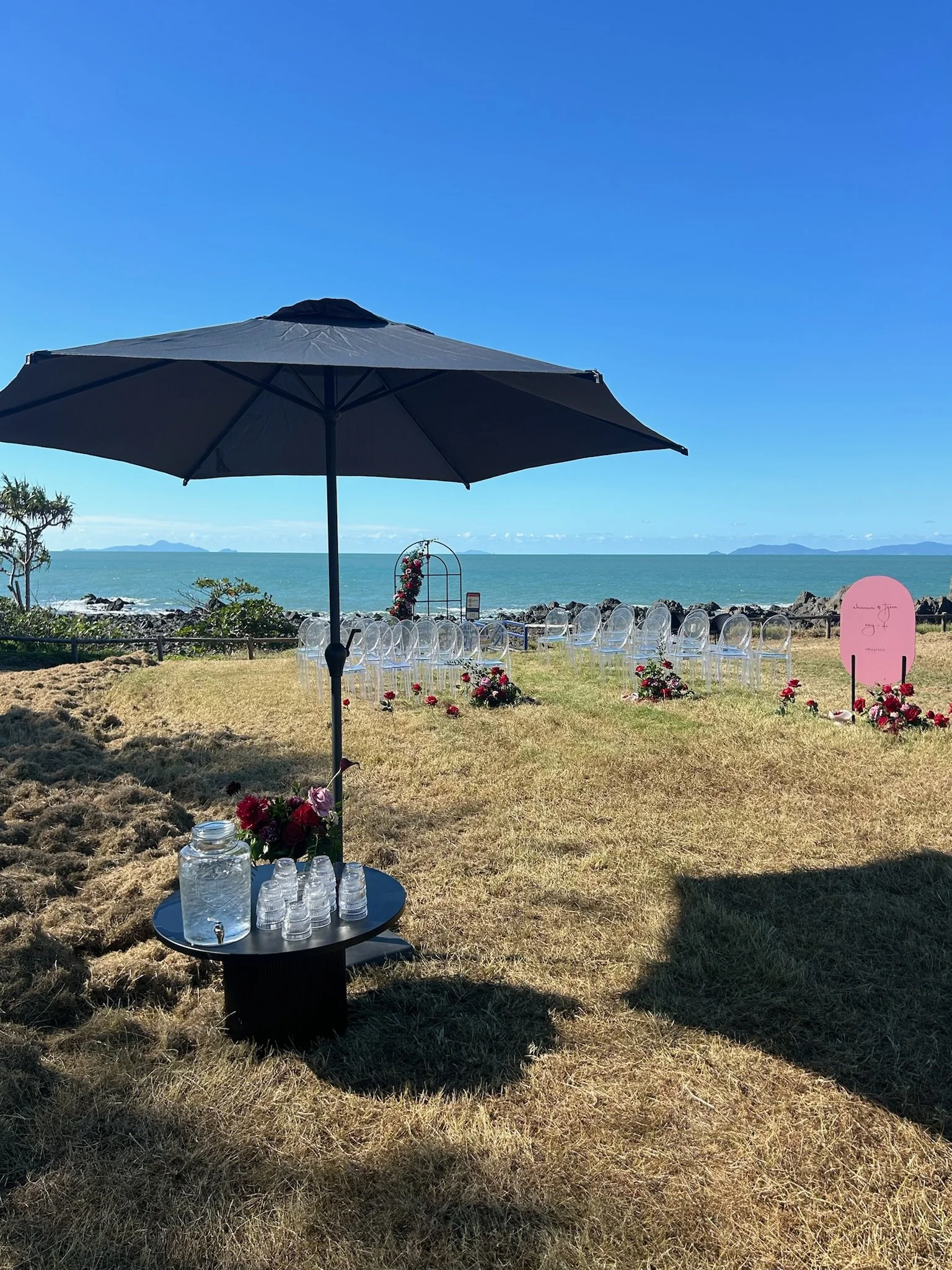 Beachside wedding setup with transparent chairs, floral arrangements, and a pink sign, under a large umbrella with the ocean and islands in the background.