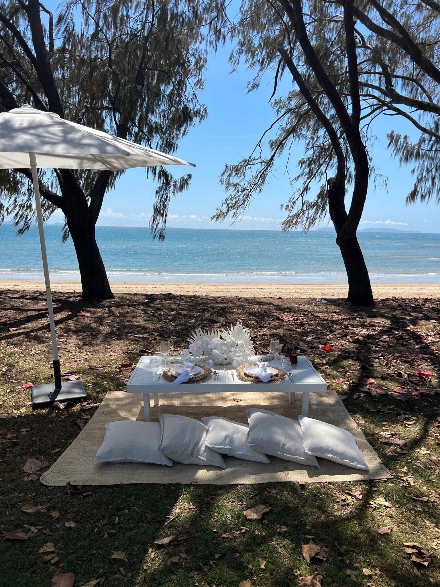 Beach setup with a white low table on a beige rug, surrounded by pillows, under trees with a view of the ocean in the background, with a white umbrella for shade and a clear blue sky.