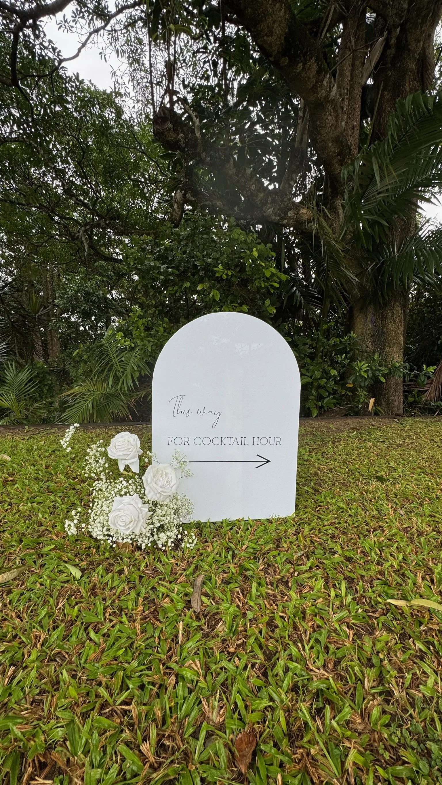 White sign with black text and arrow, decorated with white roses and baby's breath flowers, placed on green grass against a backdrop of trees and foliage, directing guests to cocktail hour.