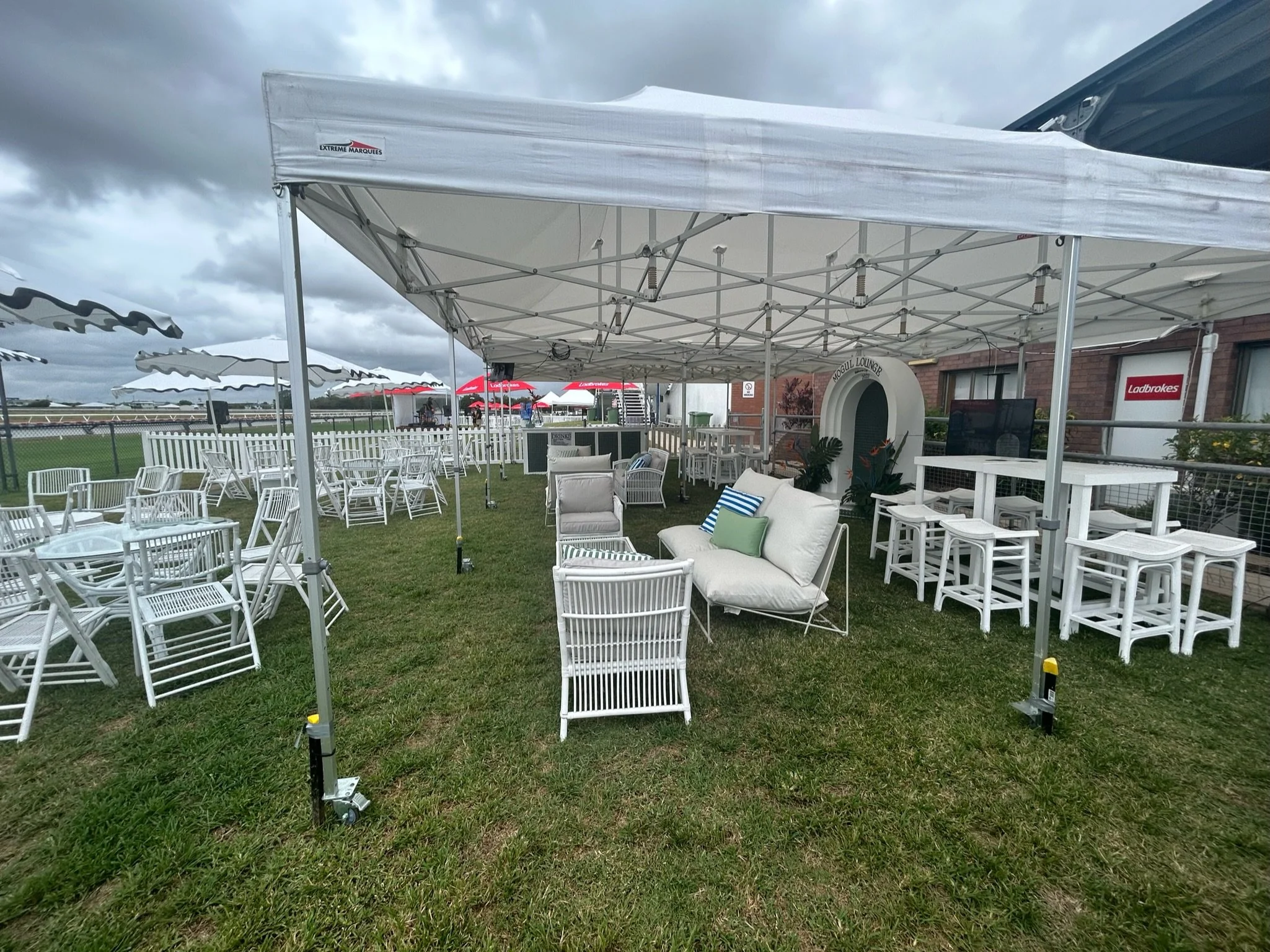 Outdoor event space with white chairs, tables, and lounge furniture under a large white canopy, on a grassy area with a cloudy sky.
