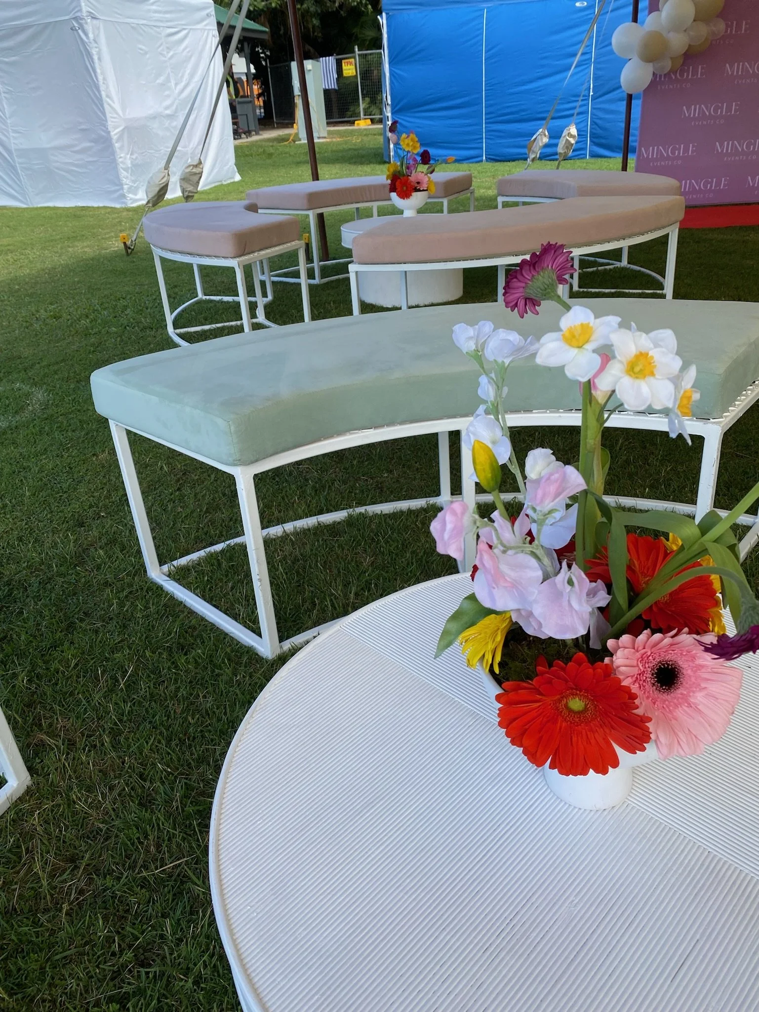 A flower arrangement with various colorful flowers, including pink, red, yellow, white, and purple, placed on a white round table outdoors. Behind it are designed outdoor chairs with cushioned seats and backrests, and balloons and tents are visible i