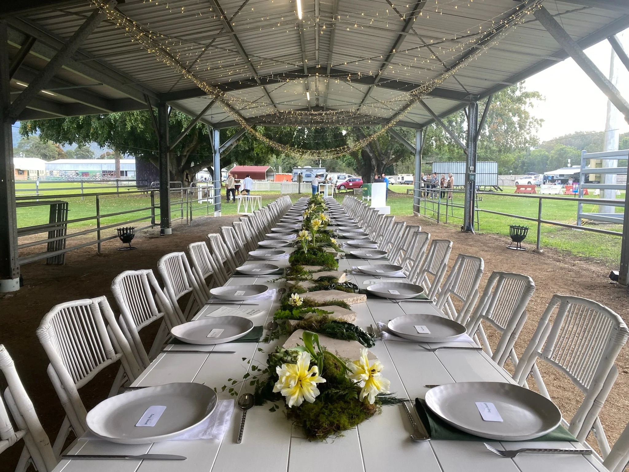 Long rectangular table set with gray plates, green napkins, and silverware under a covered outdoor shelter decorated with string lights. Floral centerpieces with white and yellow flowers run along the middle of the table. People are visible in the ba