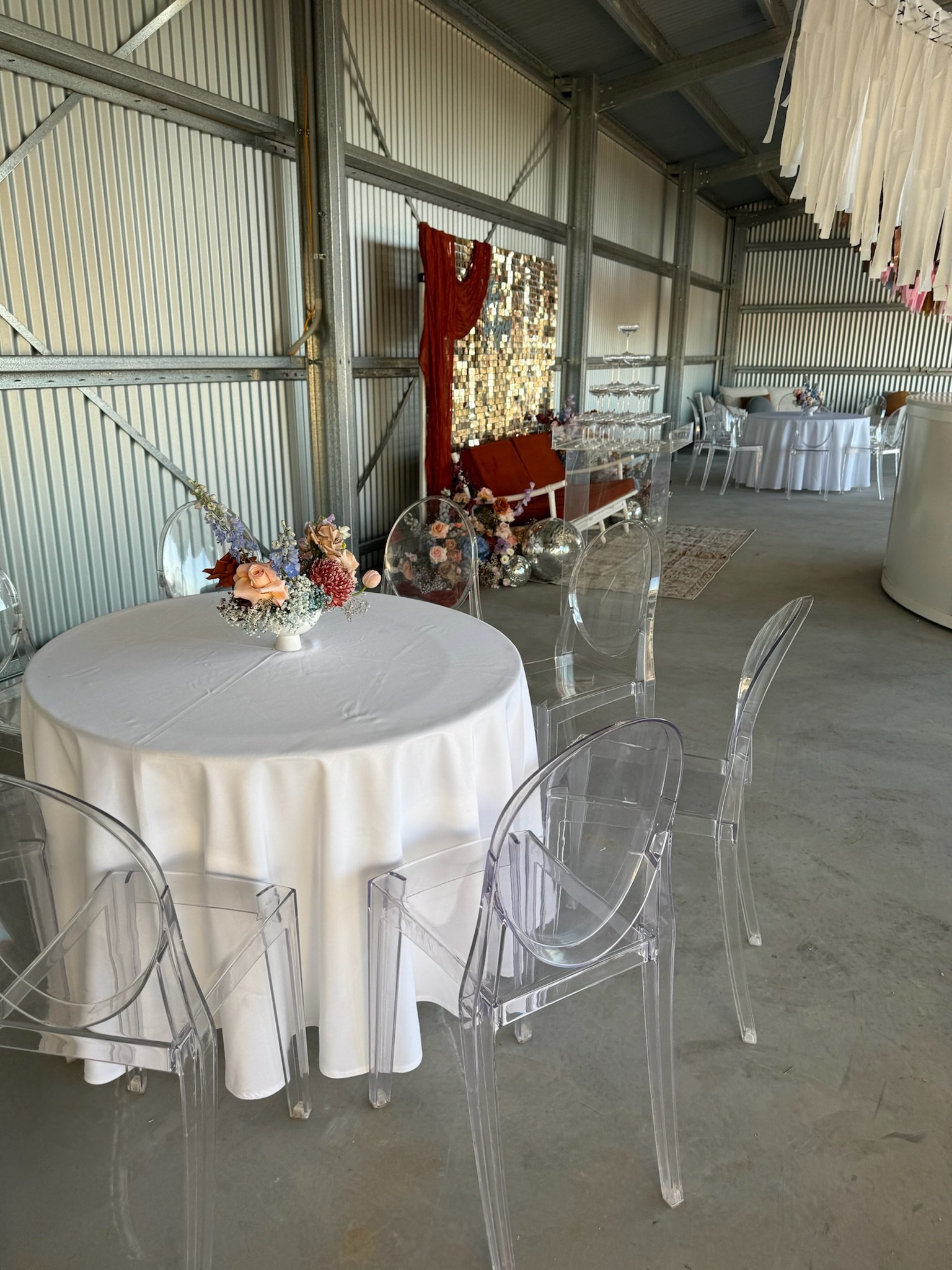 Event space with round table covered in white tablecloth, floral centerpiece, transparent ghost chairs, and decorated wall with fabric and artwork.