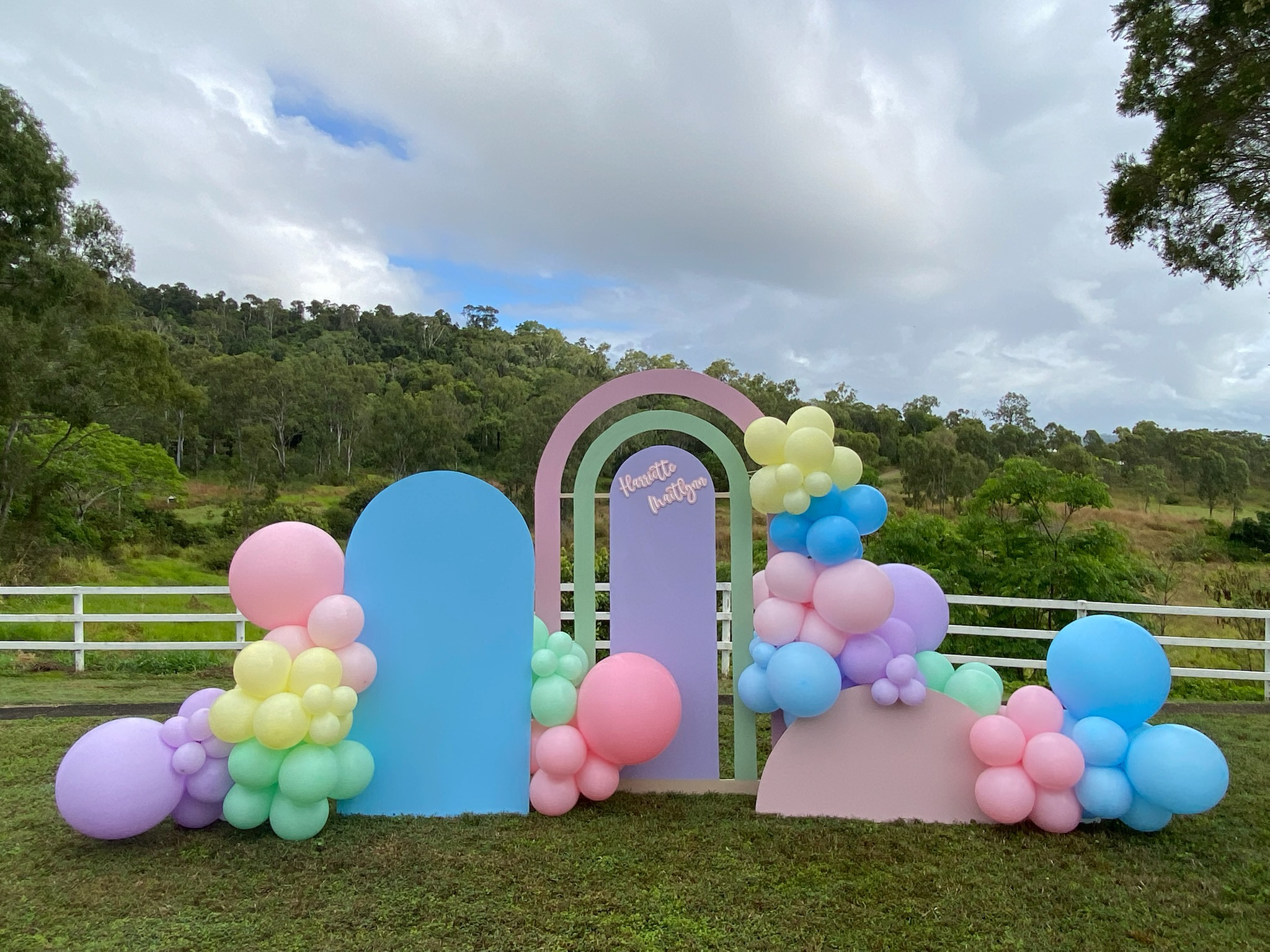 Decorative arch with pastel-colored balloons and a sign reading "Henna & More" set outdoors on grass with trees and cloudy sky in the background.