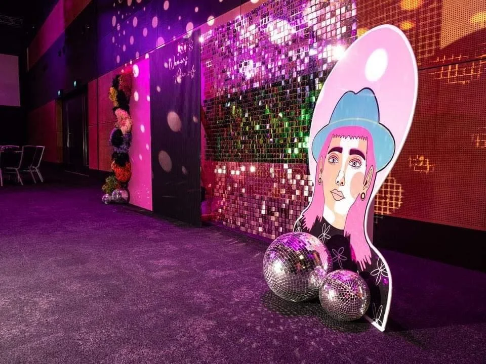 Colorful indoor party event with a large illustration of a woman with pink hair and a blue hat, surrounded by disco balls and a shiny, tiled backdrop.