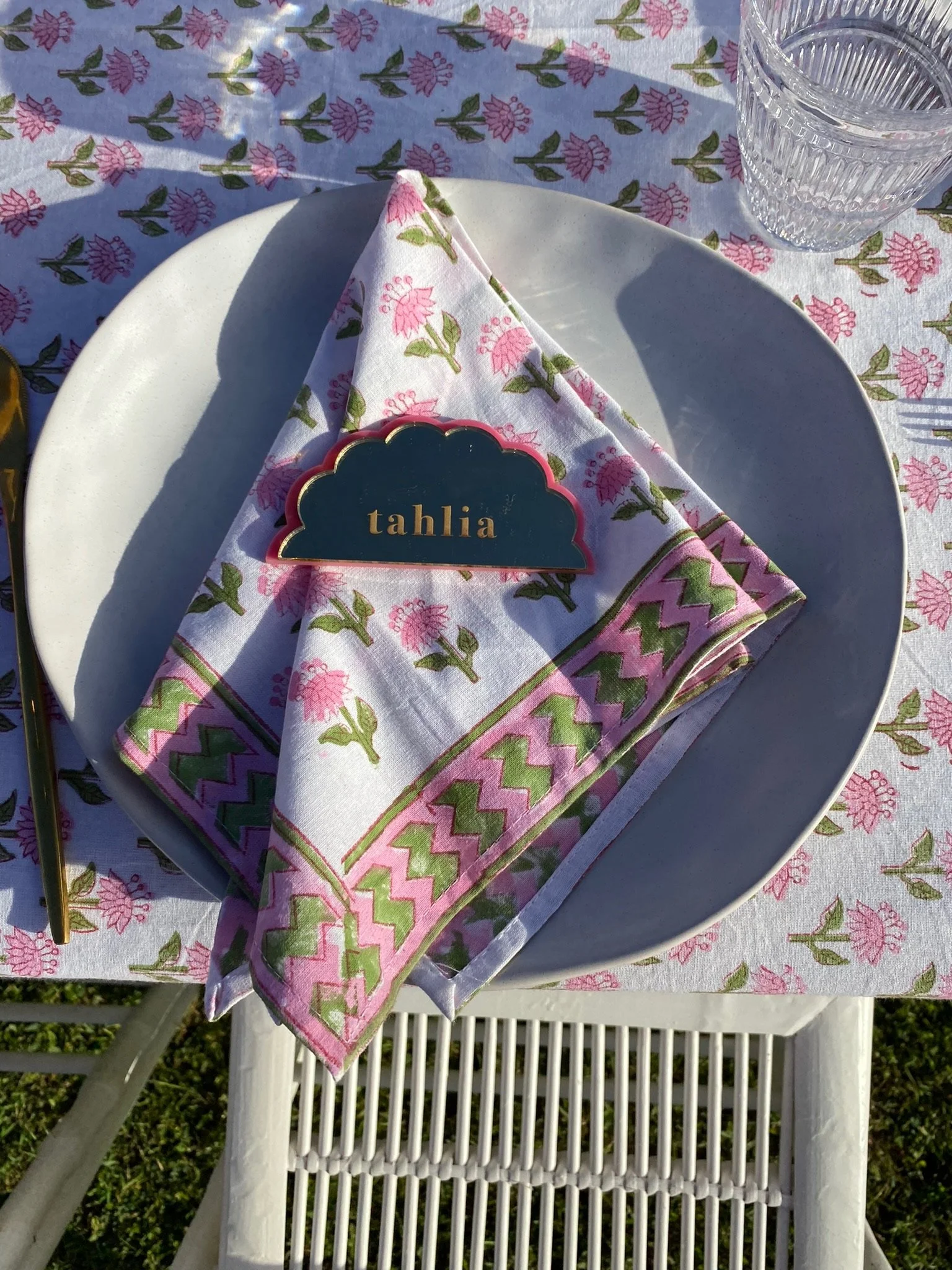 A white plate with a pink and green floral patterned napkin folded on it, with a name tag labeled 'tahlia' placed on top. The setting is outdoors on a floral tablecloth matching the napkin pattern, with two clear textured glasses in the background.