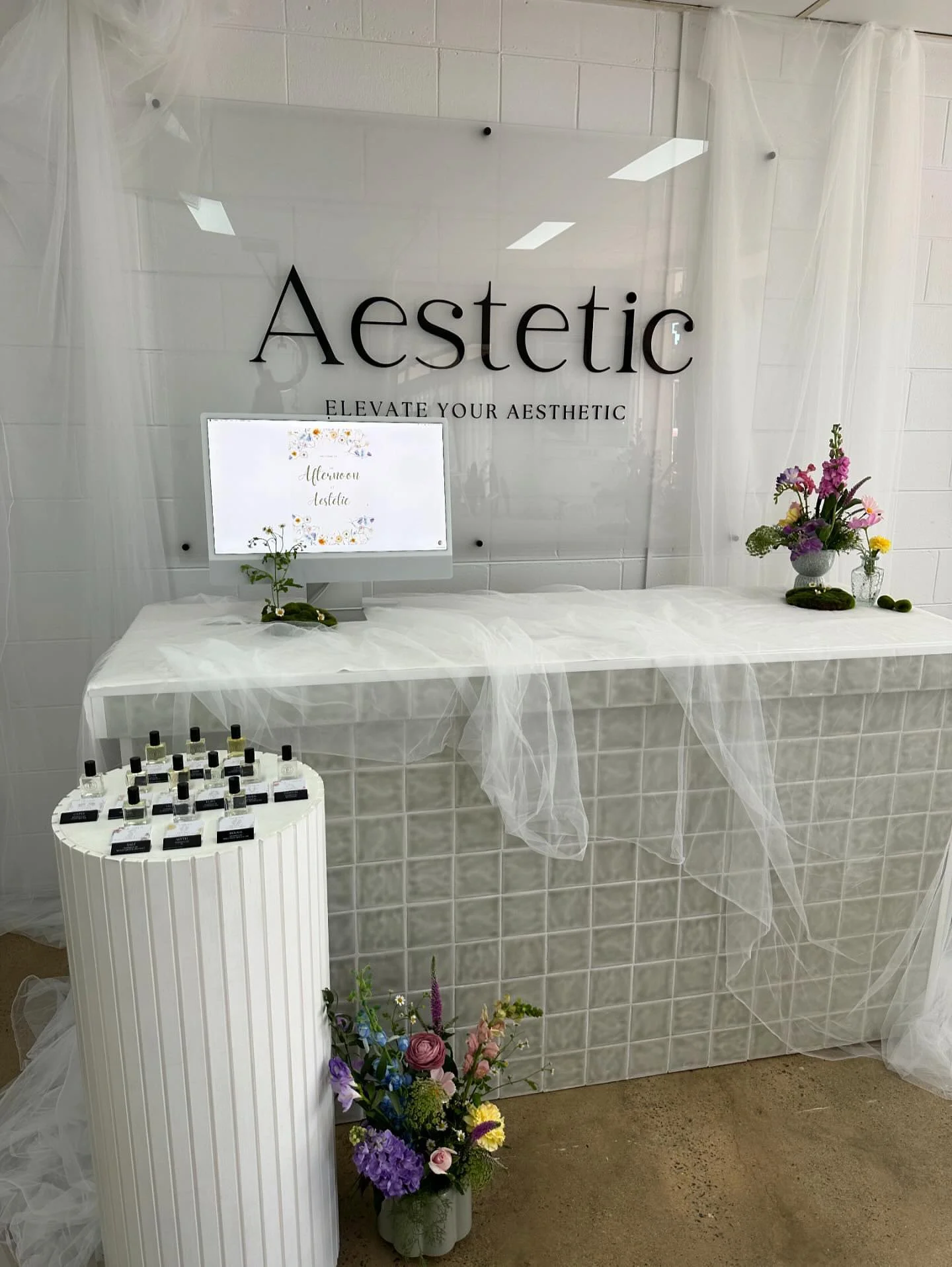 A decor setup at Aesthetic with a white-tiled front desk, flowers, and small bottles of essential oils, with a large sign on the wall that says "Aesthetic" and "Elevate your aesthetic."