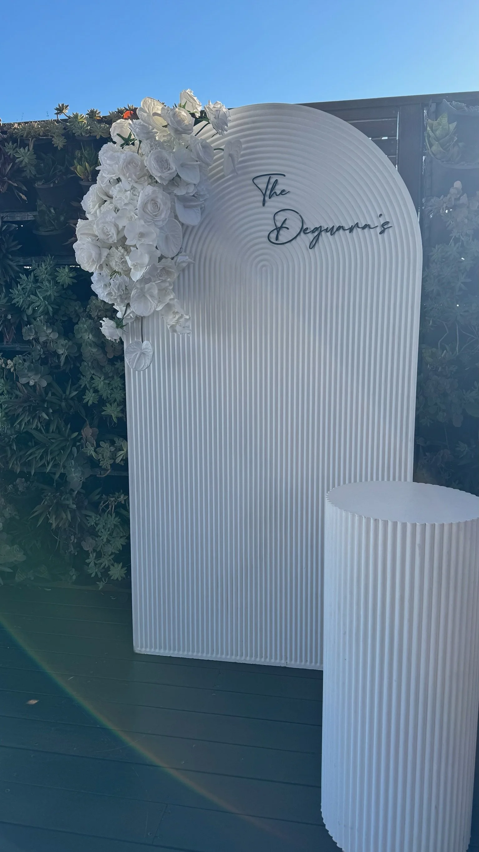 White decorative backdrop with floral arrangement and the text 'The Degunaro's' at the top right corner, next to a large white cylindrical pedestal, set outdoors under a clear blue sky.