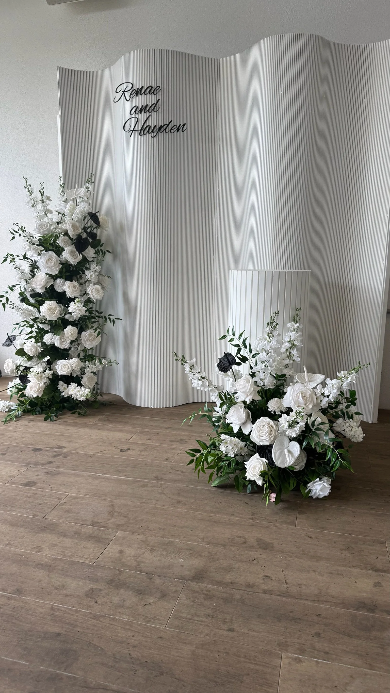 Wedding backdrop with white floral arrangements and a white textured screen with the names "Renae and Hayden" written on it.