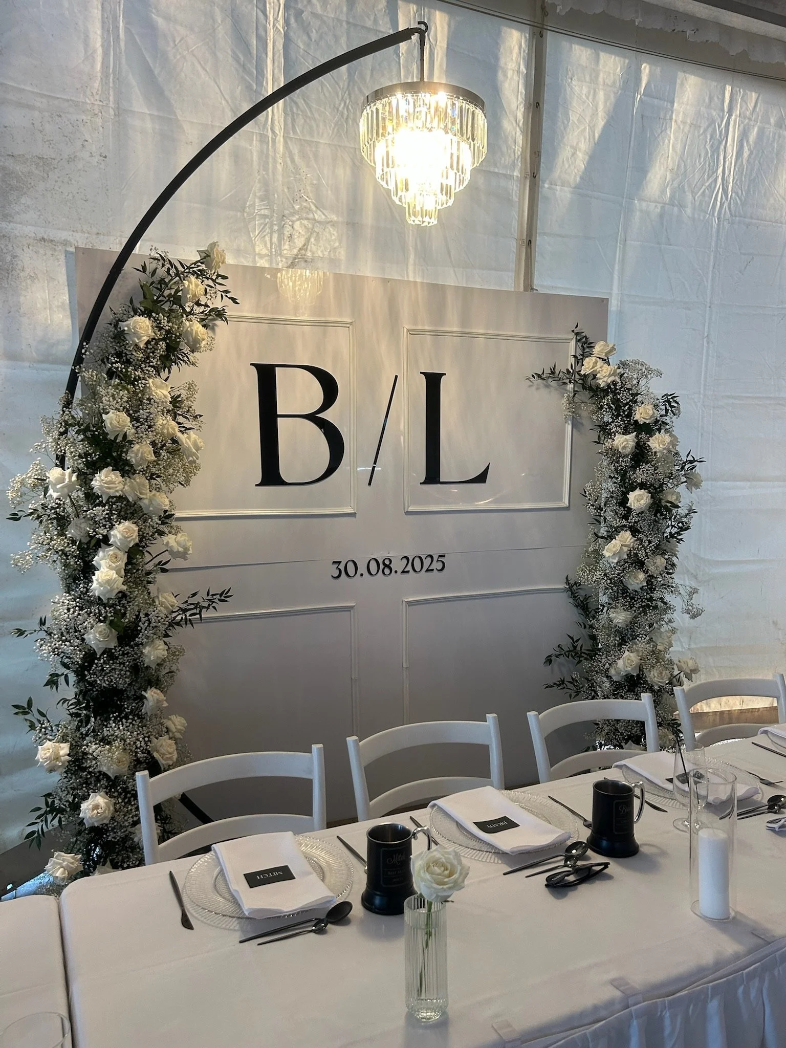Wedding reception setup with a white table, chairs, flower arrangements, and a decorative backdrop displaying initials 'B/L' and the date 30.08.2025. There is a chandelier overhead and a large floral arch.