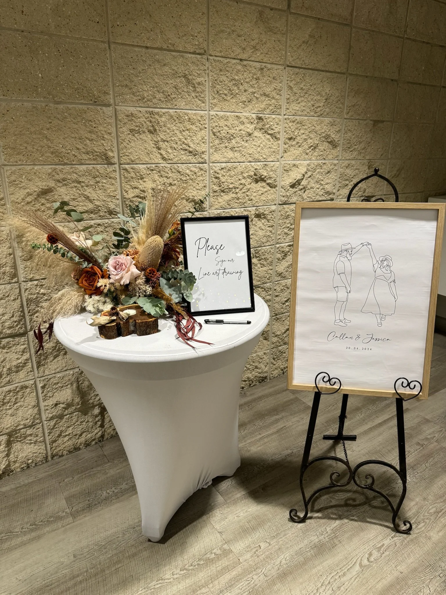Table with floral arrangement, sign-in sheet, and a framed artwork of a couple with the names Collin & Jessica and the date 20.04.2024, set against a brick wall at a wedding or event reception.