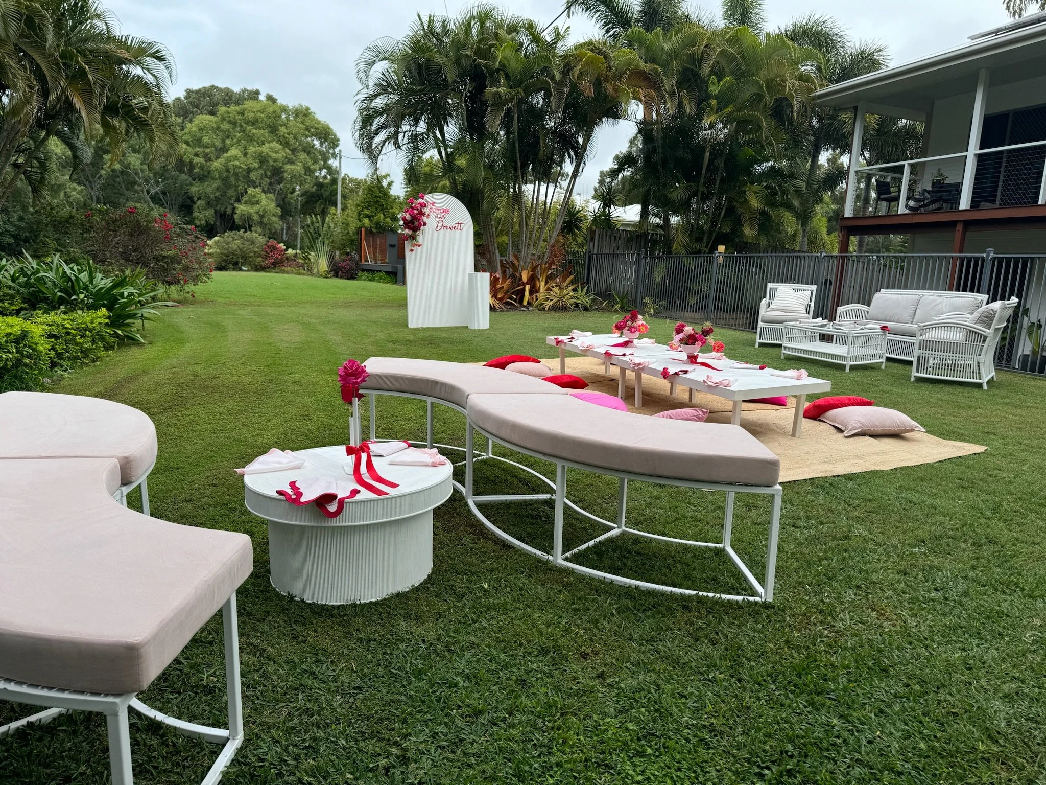 Outdoor garden setting decorated for a celebration with low tables, pillows, and floral arrangements, surrounded by greenery and patio furniture.
