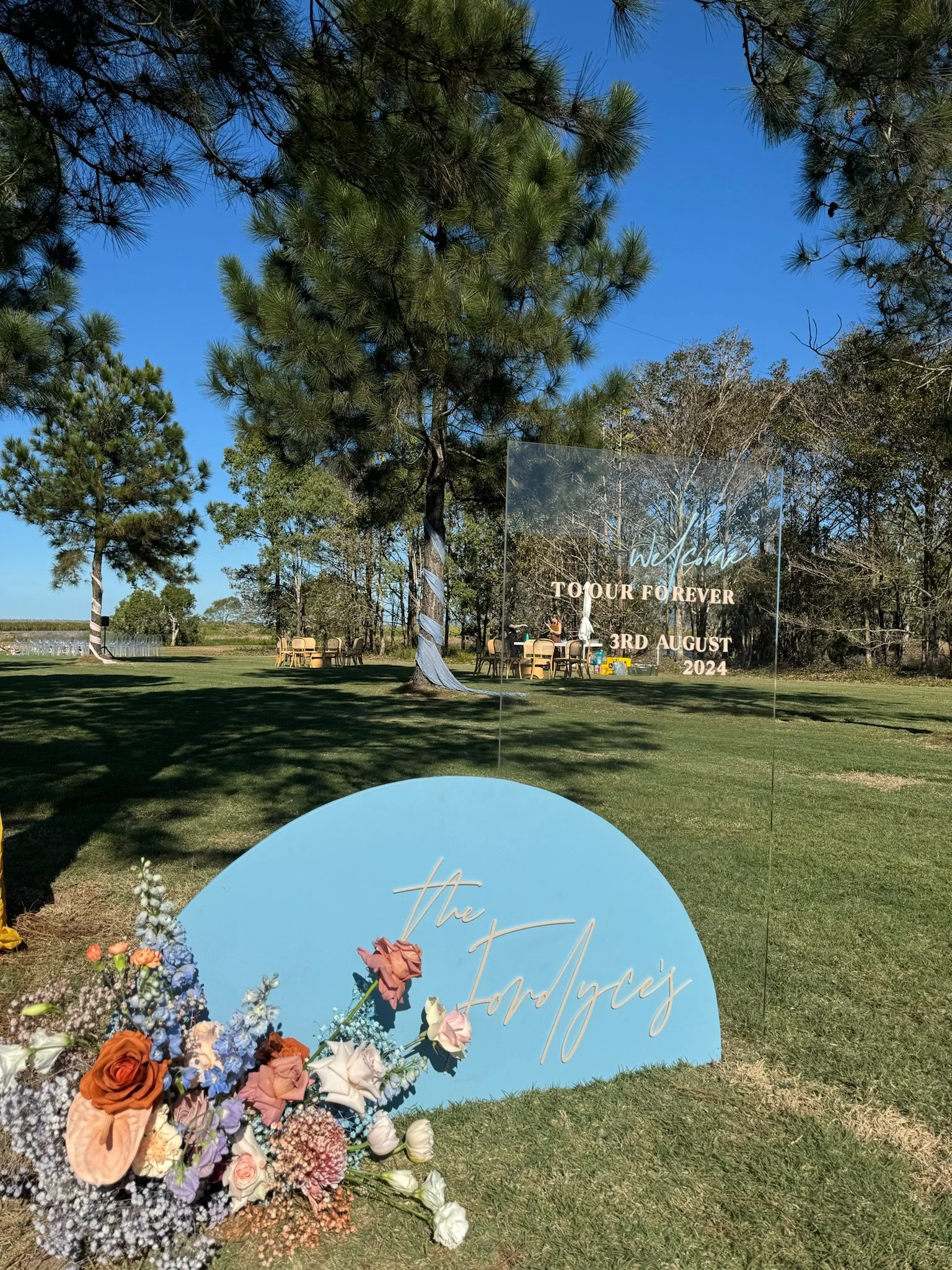 Outdoor wedding or event setup with a blue circular sign and floral arrangements in front. The sign reads 'The Fordyce's,' with the larger transparent sign saying 'Welcome TO OUR FOREVER 3RD AUGUST 2024' in the background. There are tables and chairs