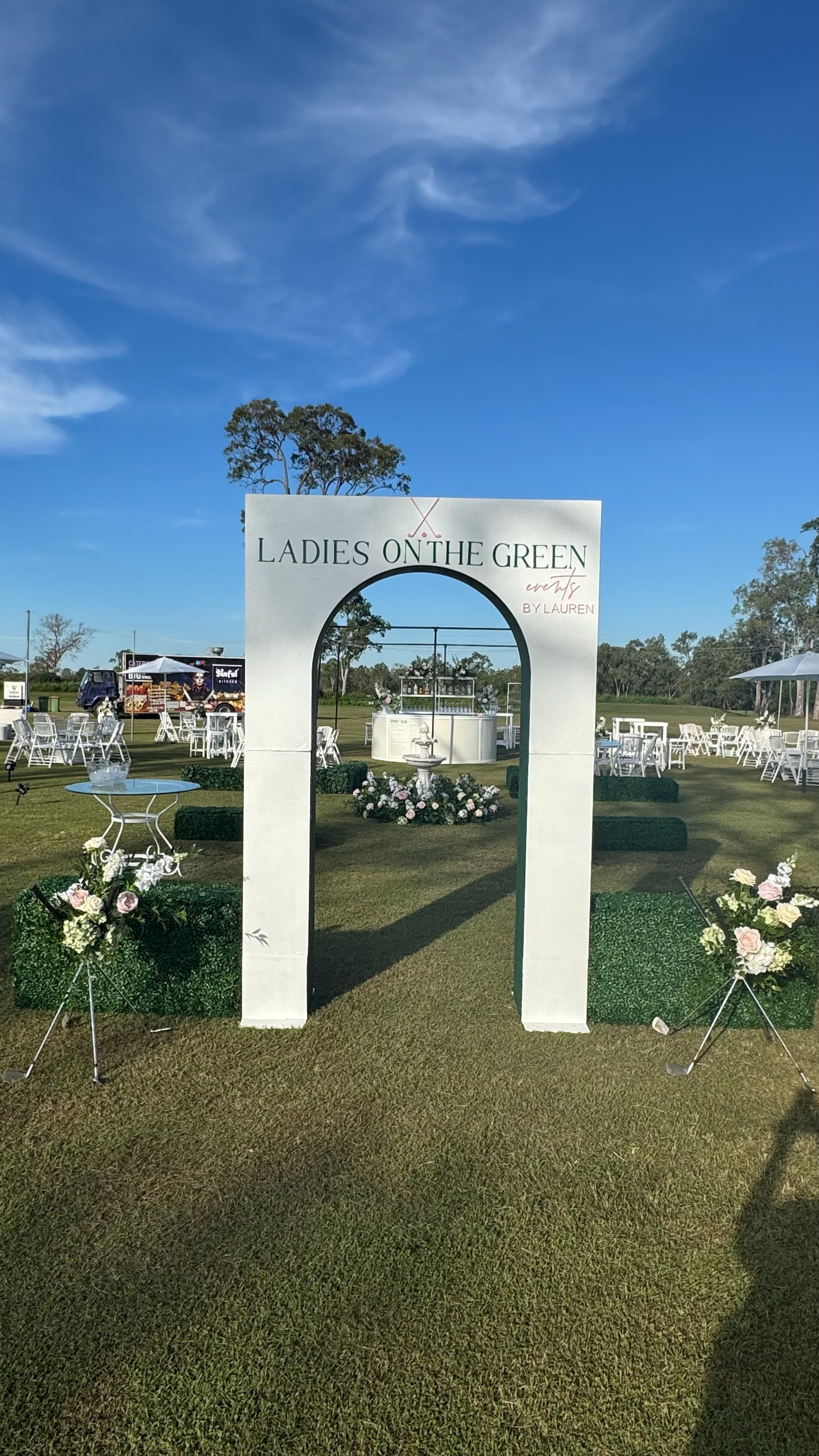 Outdoor event setup with white arch entrance labeled "Ladies on the Green", decorated with flowers, leading to a white bar and seating area with white tables and chairs, under a blue sky.