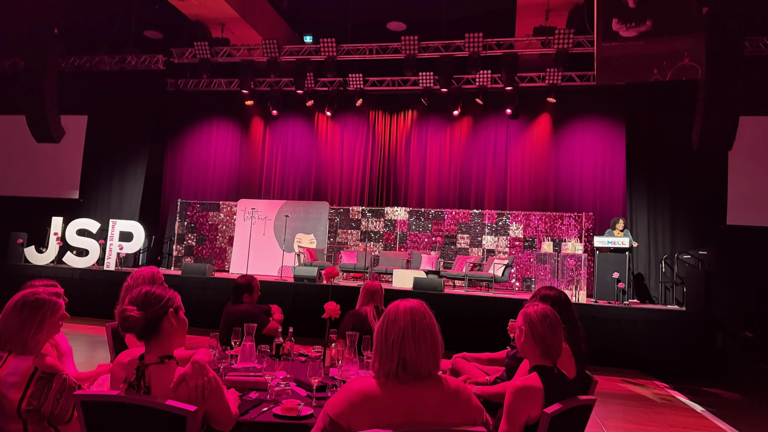 Stage decorated with pink curtains and seating area, with a woman speaking at a podium on the right, audience members seated at tables in the foreground, large illuminated letters 'JSP' on the left, and pink lighting throughout.
