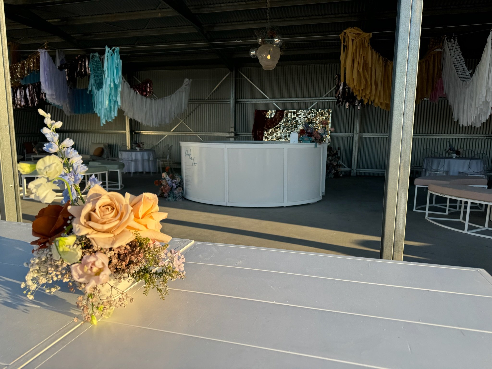 Indoor event space decorated with pastel-colored tassel garlands, floral arrangements, and white tables, with a curved white bar in the background.