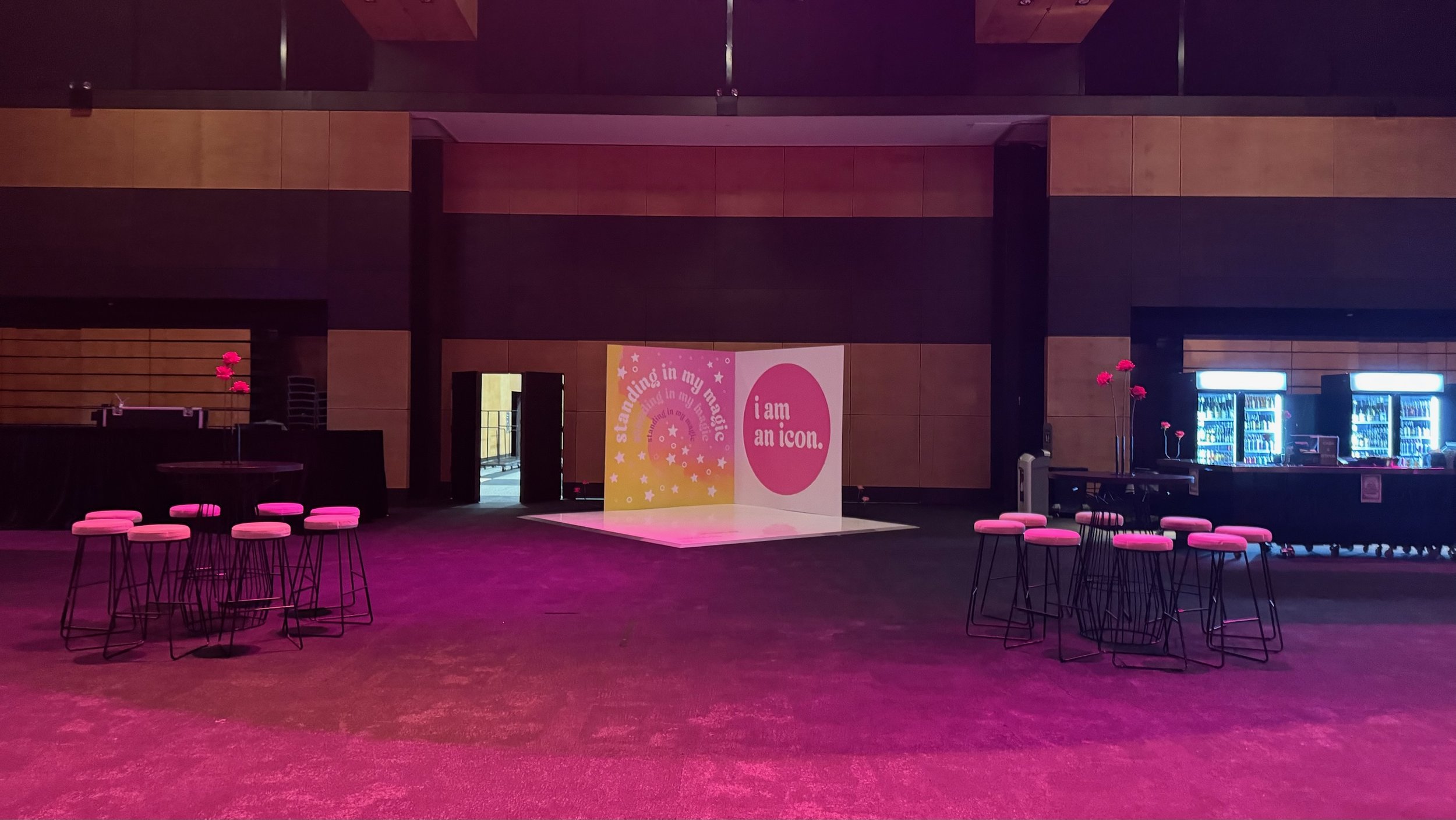 Empty event space with pink lighting, round tables with pink chairs, large pink flower centerpieces, and a backdrop with the text 'standing in my magic' and 'i am an icon' behind an open area.