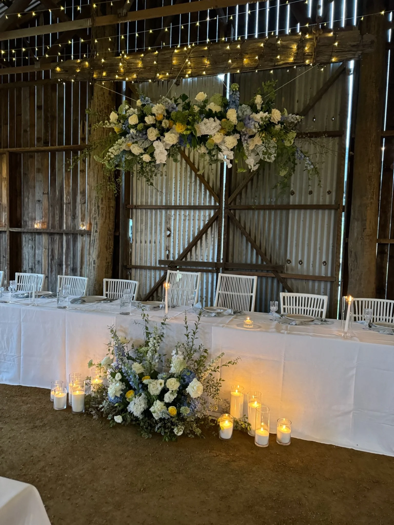 A wedding reception table set up inside a rustic barn with a white tablecloth, glassware, and place settings. A large floral arrangement with white, yellow, and blue flowers is on the floor in front of the table, and smaller candles and floral arrang